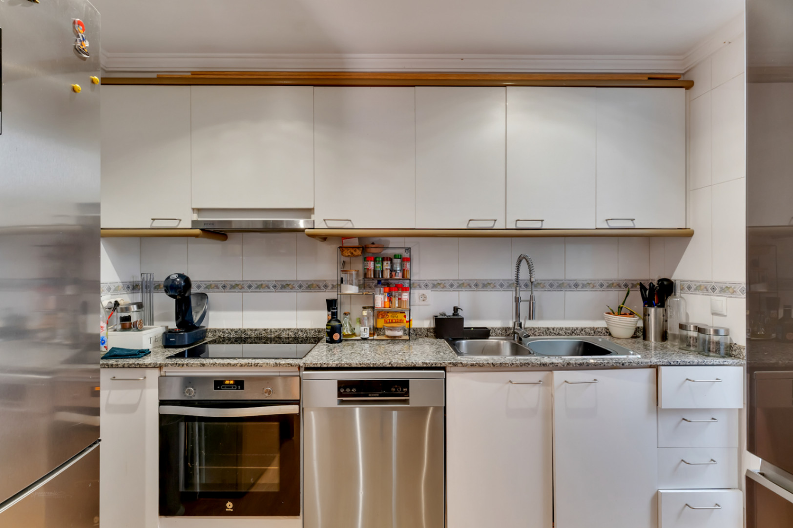Cuisine fonctionnelle avec plan de travail en granit, meubles blancs et électroménagers en inox. Équipée d'une plaque vitrocéramique et d'un lave-vaisselle intégré.