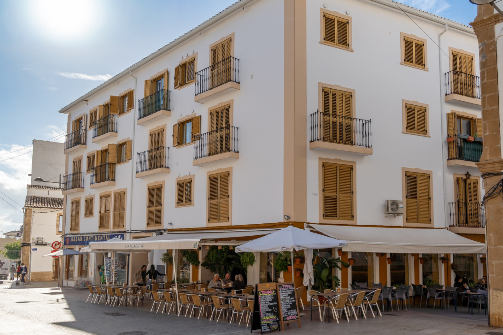 Façade traditionnelle du centre historique avec volets en bois ocre, balcons en fer forgé et terrasse de café au rez-de-chaussée d'une zone piétonne.