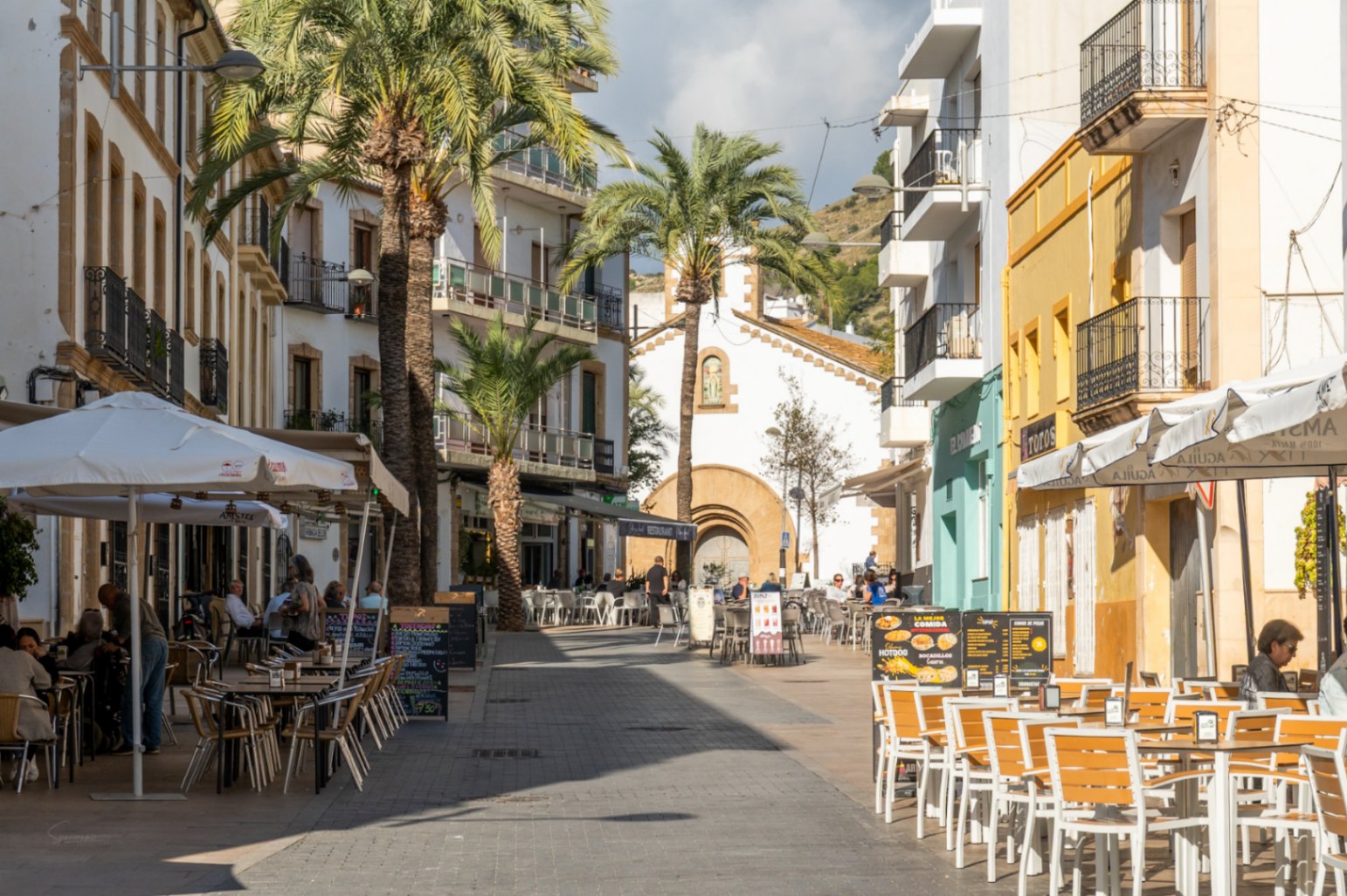 Rue piétonne du centre historique de Jávea avec terrasses de restaurants, palmiers et façades traditionnelles sous une lumière naturelle.