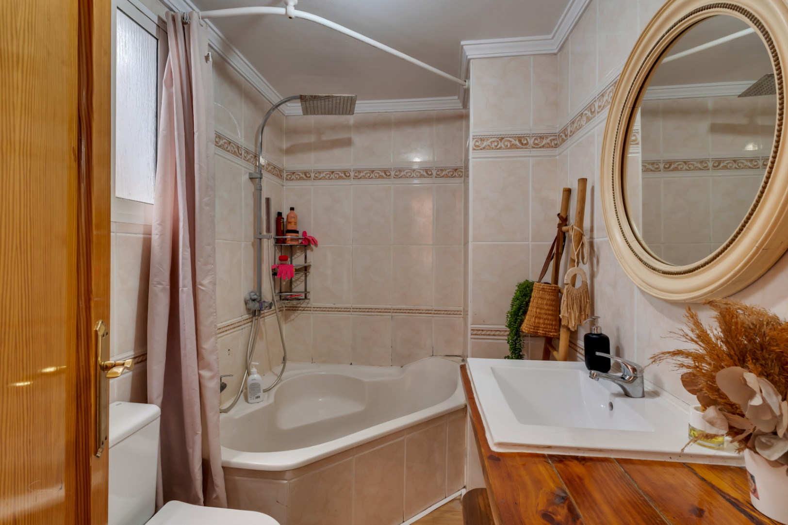Salle de bain complète avec baignoire d'angle, vasque sur plan de travail en bois rustique et miroir circulaire décoratif. Murs carrelés avec frise classique.