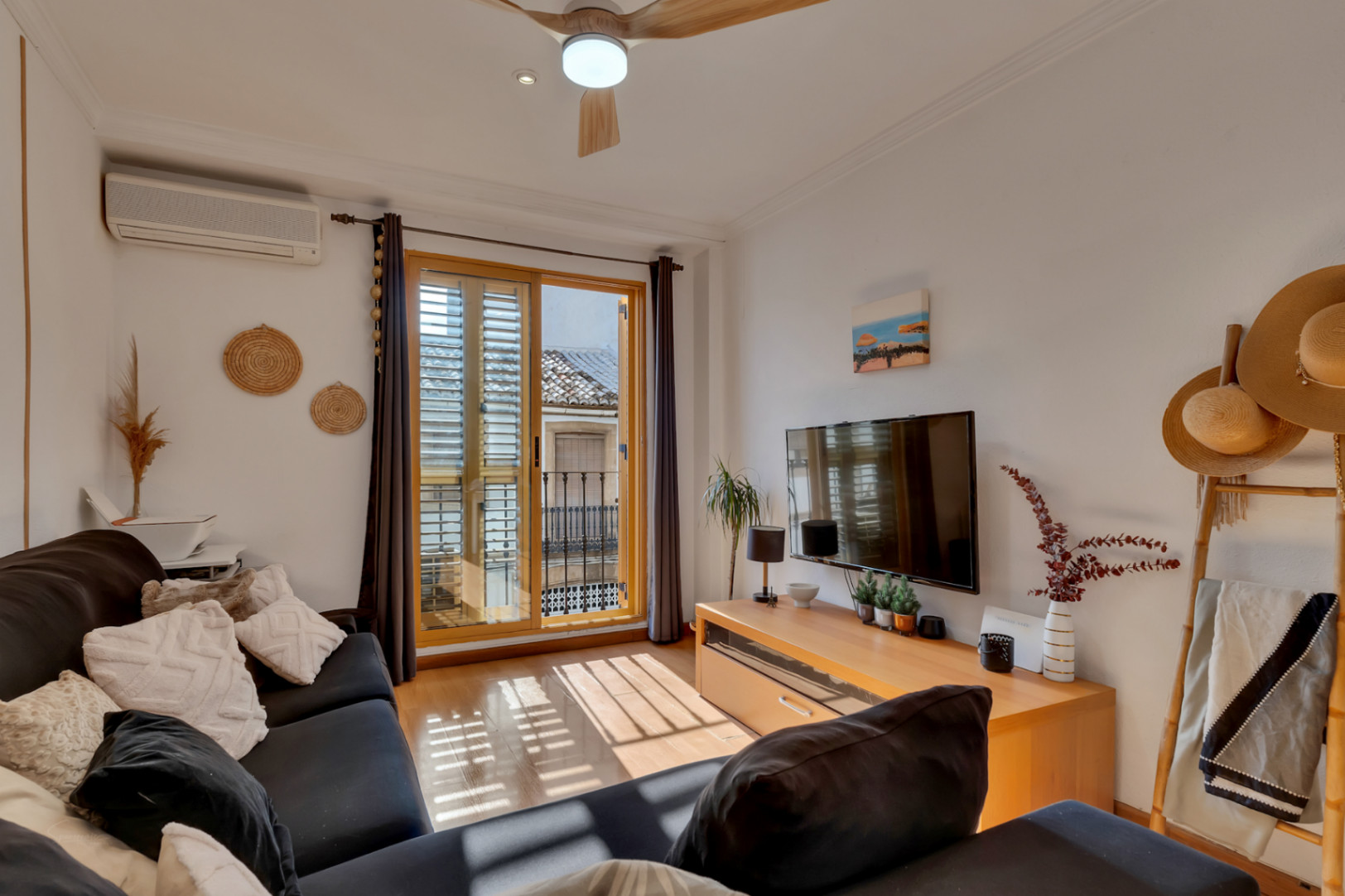 Salon lumineux avec parquet, climatisation et accès à un balcon avec volets en bois. L'espace comprend un ventilateur de plafond et un meuble TV en bois naturel.