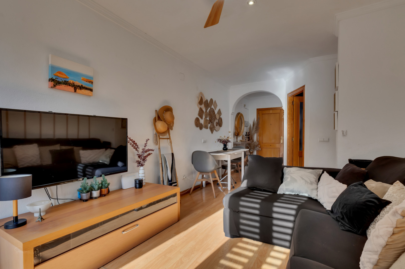 Salon-salle à manger lumineux avec parquet stratifié, canapé d'angle et coin repas. On remarque le meuble TV en bois et l'ouverture en arche vers l'entrée de l'appartement.