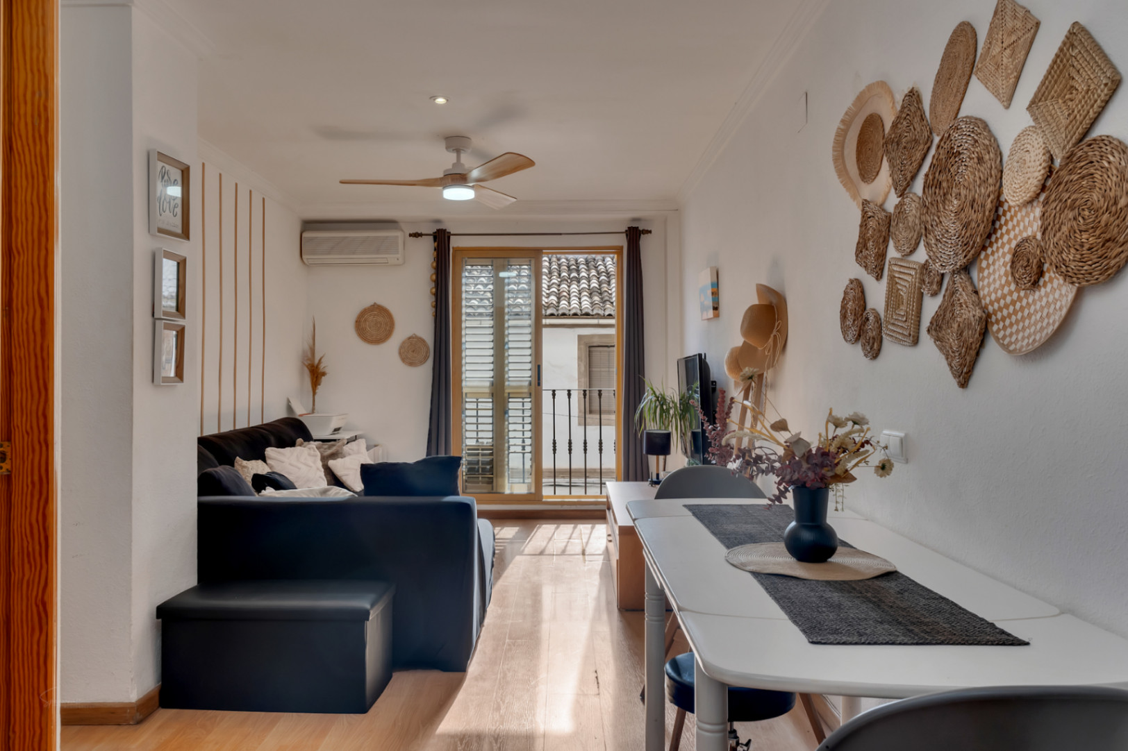 Salon-salle à manger lumineux avec parquet, ventilateur de plafond et accès au balcon. La décoration méditerranéenne intègre des éléments artisanaux et des tons neutres.