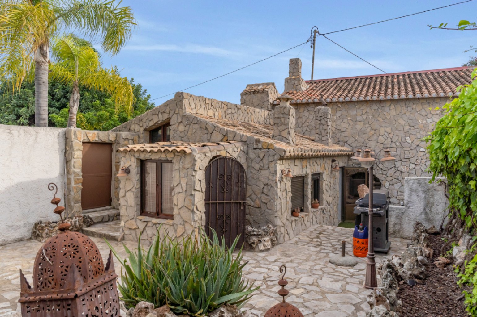 Appartement d'invités indépendant avec façades en pierre naturelle, toiture en tuiles et terrasse pavée équipée d'un barbecue et de plantes méditerranéennes.