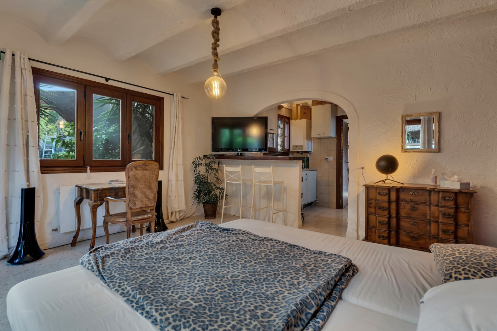 Chambre avec bureau en bois, plafond texturé et arche ouvrant sur une kitchenette fonctionnelle équipée d'un comptoir et de tabourets.