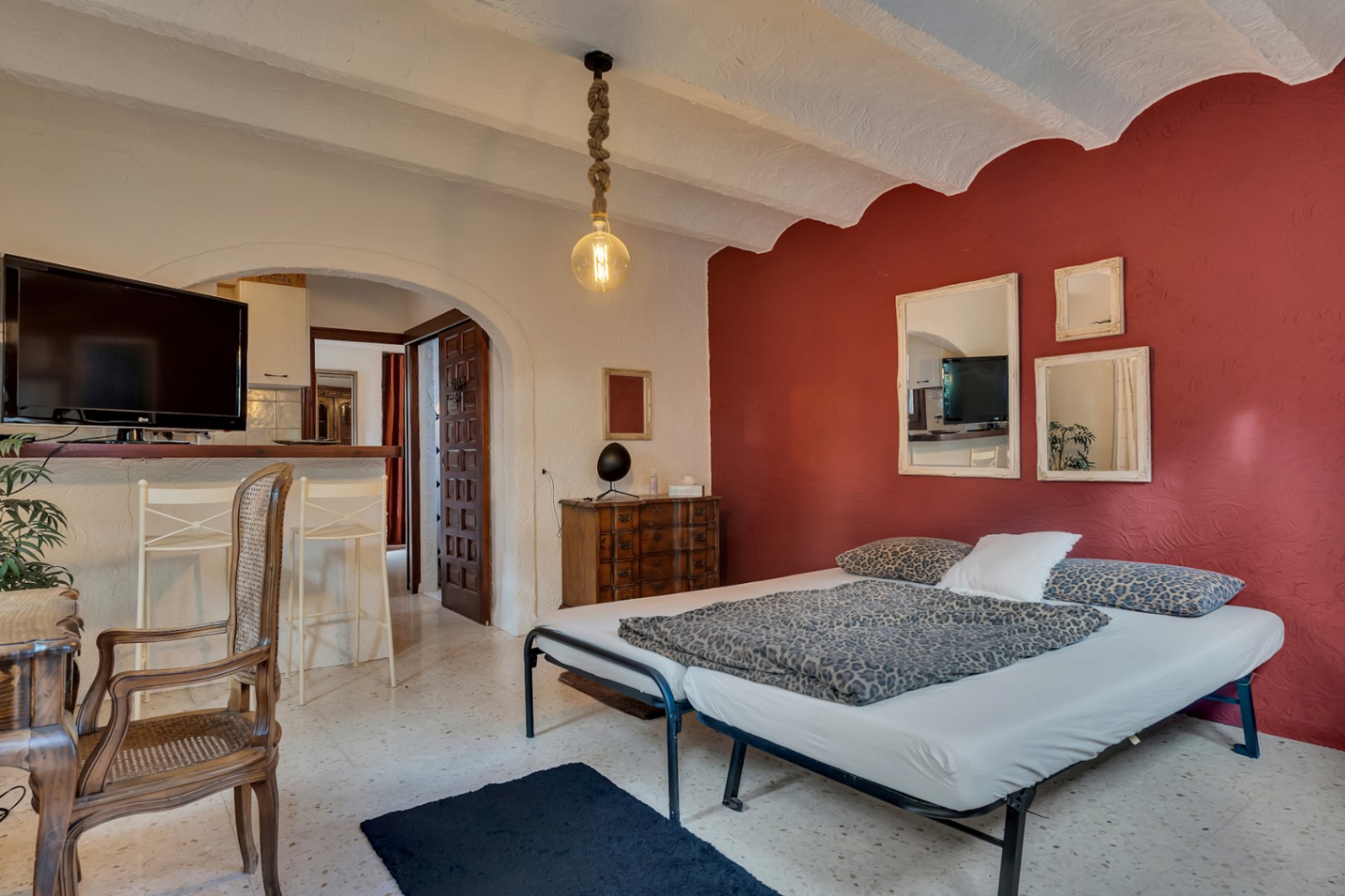 Chambre avec mur d'accent rouge, plafonds voûtés et sol en terrazzo. Elle dispose d'une ouverture voûtée vers le séjour et d'une suspension en corde.