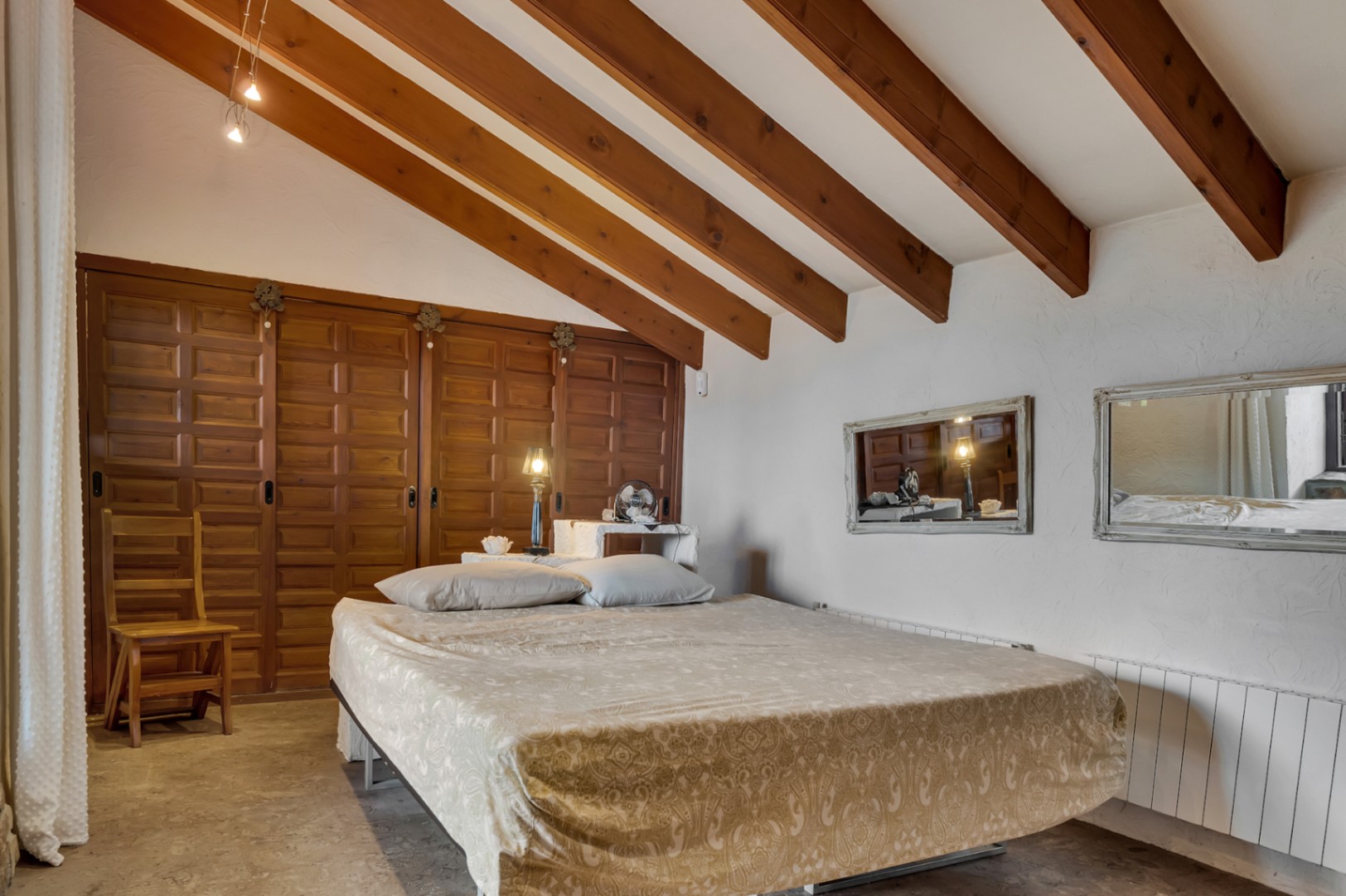 Chambre avec plafond incliné aux poutres en bois apparentes, grands placards intégrés en bois et murs blancs à la finition texturée.