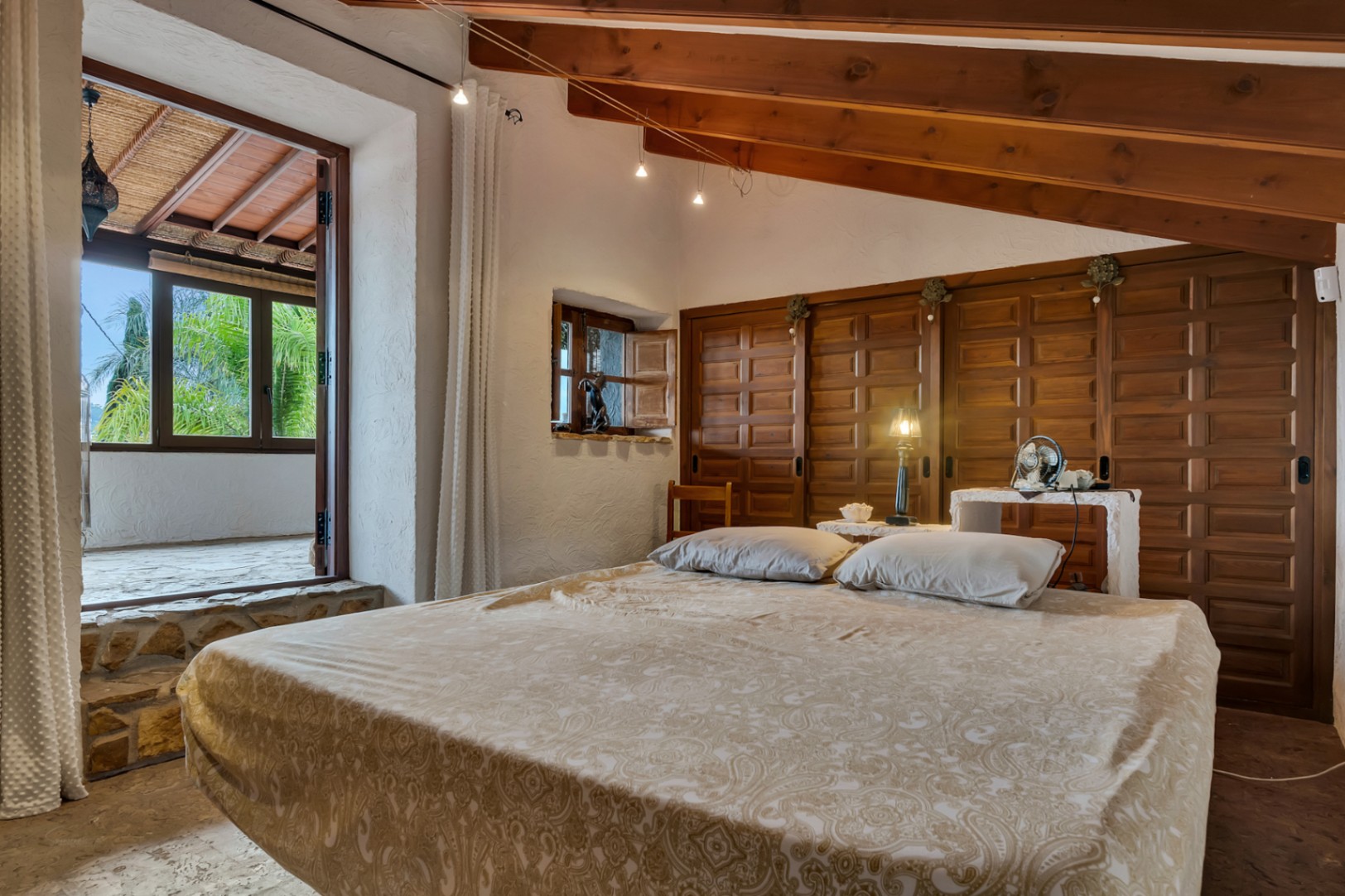 Chambre avec plafonds à poutres apparentes, placards intégrés en bois et accès direct à une terrasse couverte par un seuil en pierre naturelle.