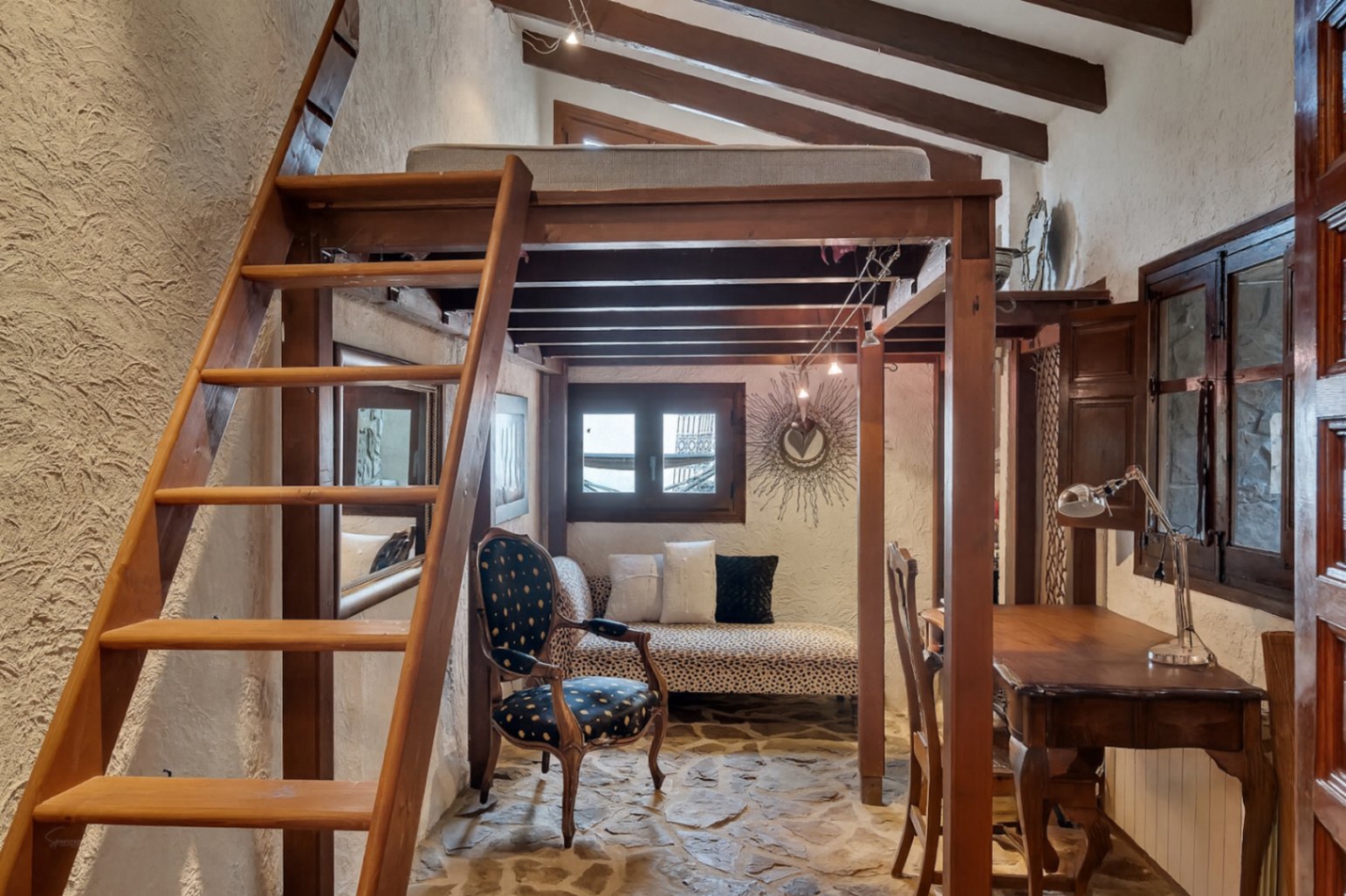 Chambre rustique avec mezzanine en bois, poutres apparentes et sol en pierre naturelle. L'espace dispose d'un coin bureau et d'un salon sous la structure.