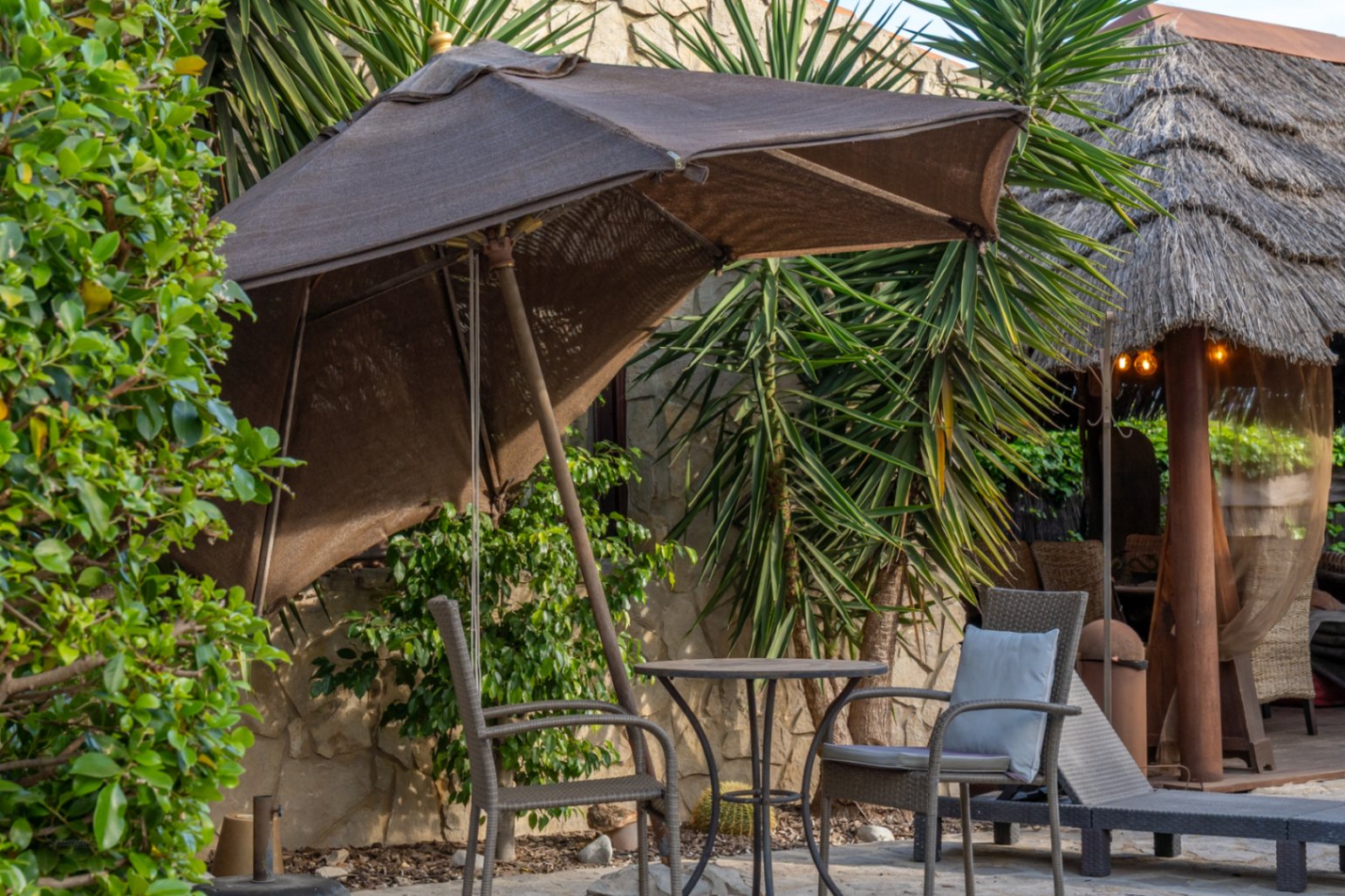 Coin salon extérieur avec parasol, table de bistrot et chaises en osier, situé devant un mur en pierre naturelle et une pergola balinaise au toit de chaume.