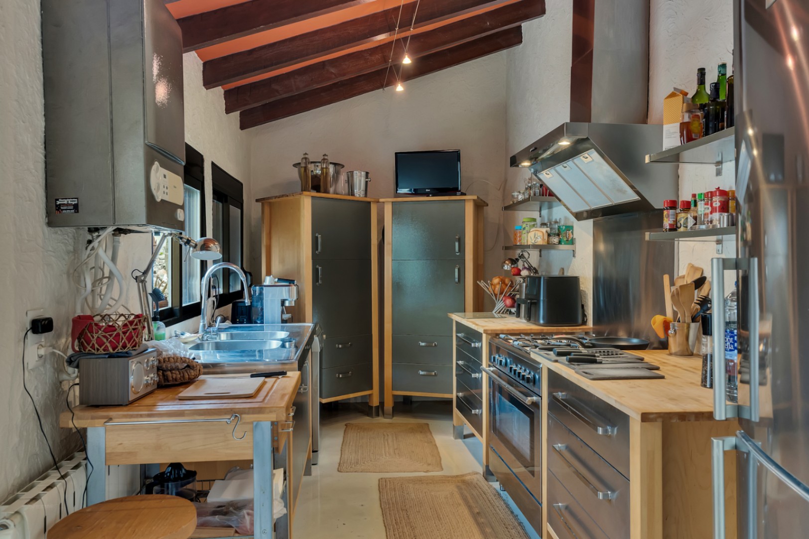 Cuisine de style industriel avec poutres en bois apparentes, plans de travail en bois massif et appareils en acier inoxydable sous un plafond incliné.