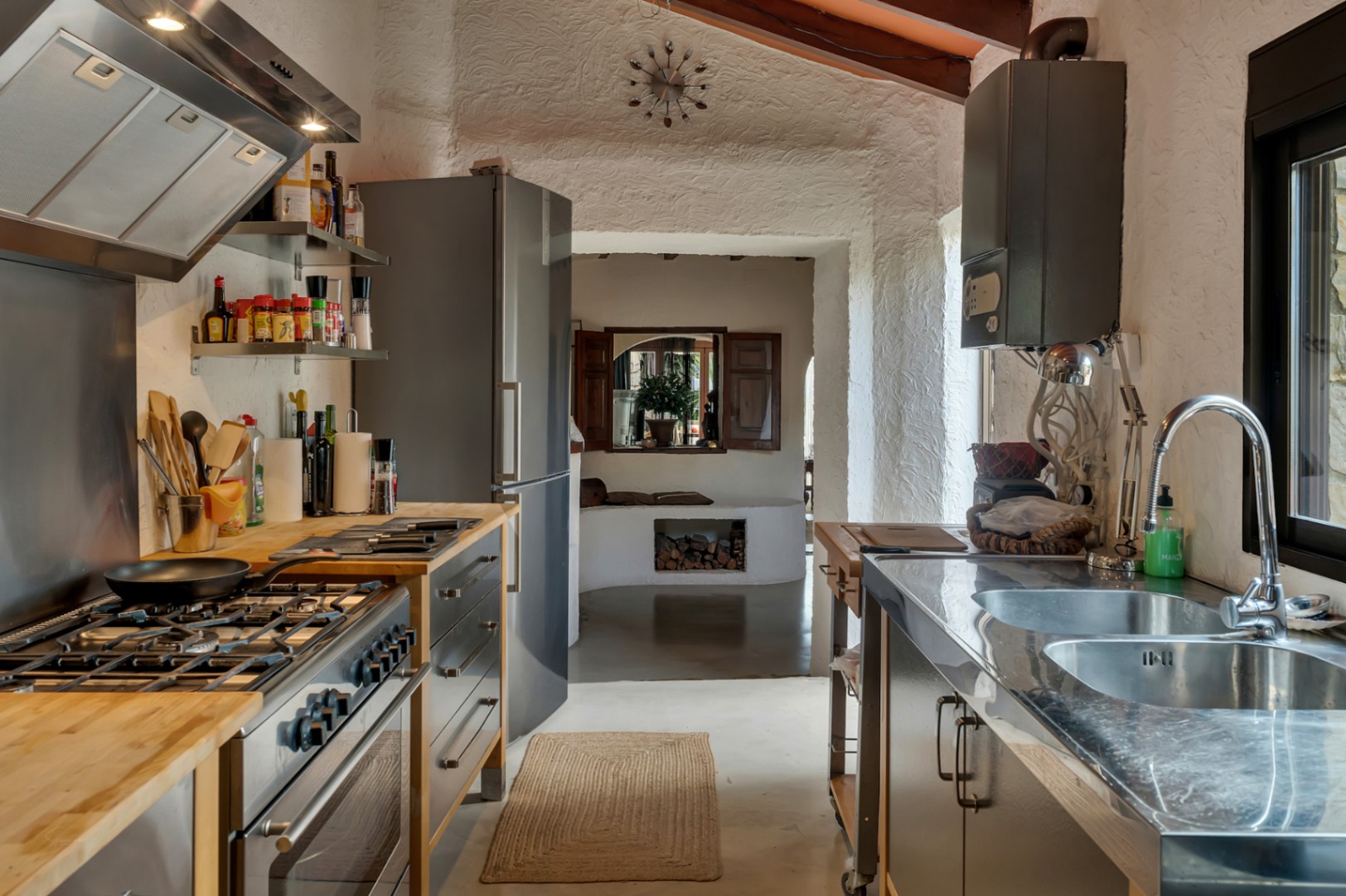 Cuisine rustique moderne avec plans de travail en bois et inox, murs texturés blancs et vue sur une pièce de vie avec cheminée à bois.