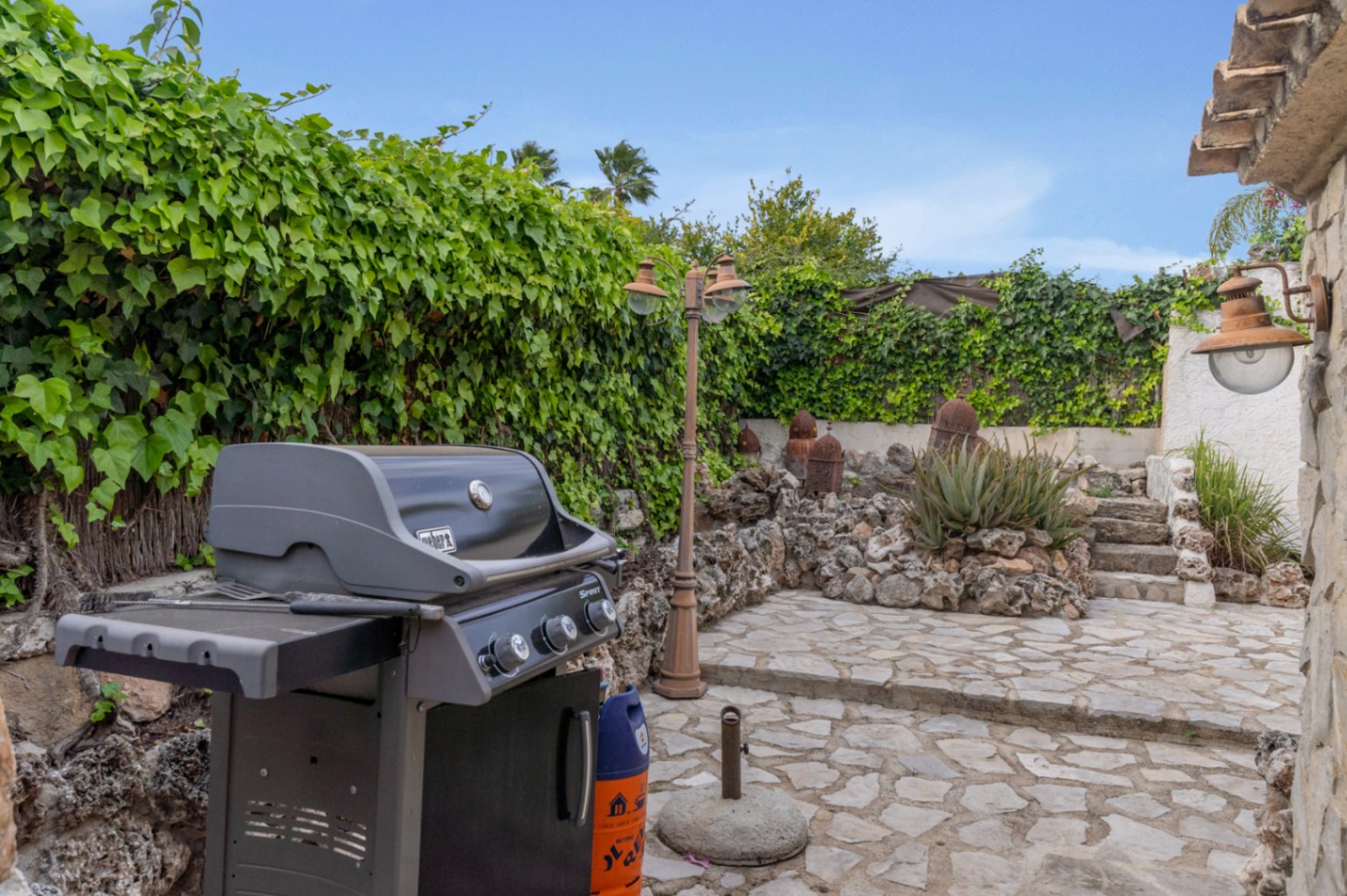 Espace barbecue extérieur avec grill à gaz Weber, sol en pierre naturelle et murs recouverts de lierre au sein d'un jardin méditerranéen soigné.