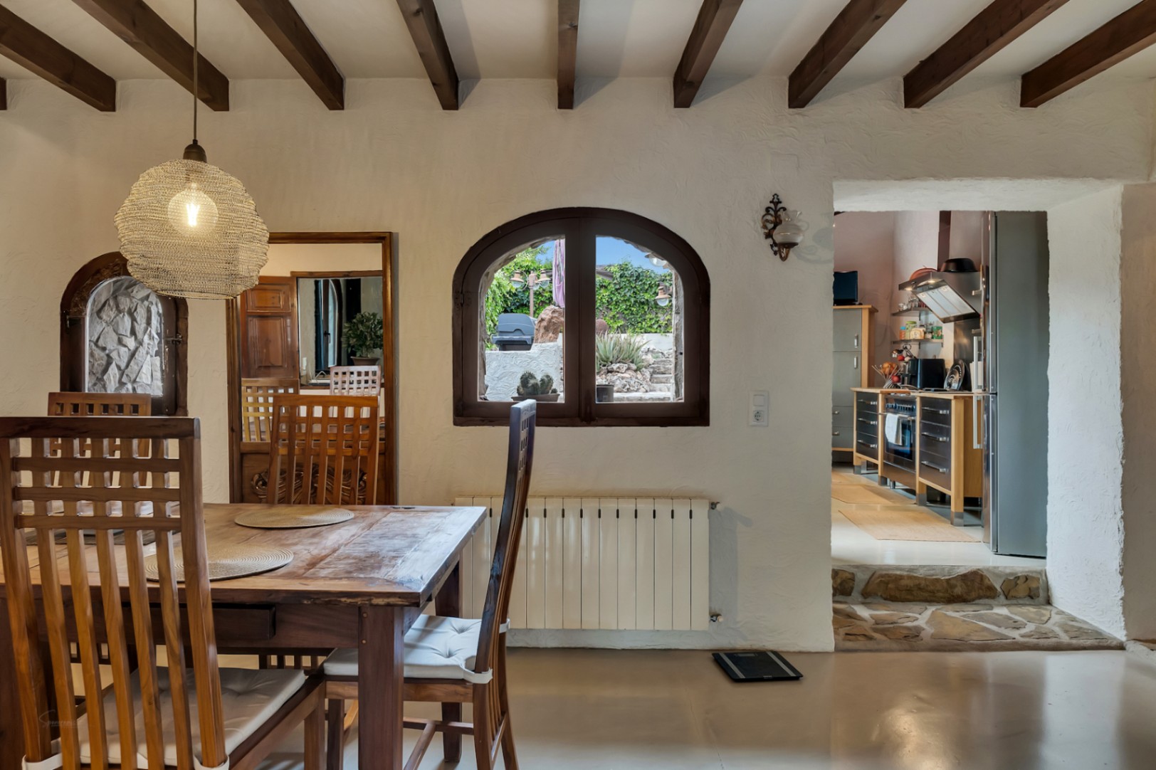 Espace dînatoire avec poutres en bois, fenêtre cintrée vue jardin et accès à la cuisine moderne par une marche en pierre.