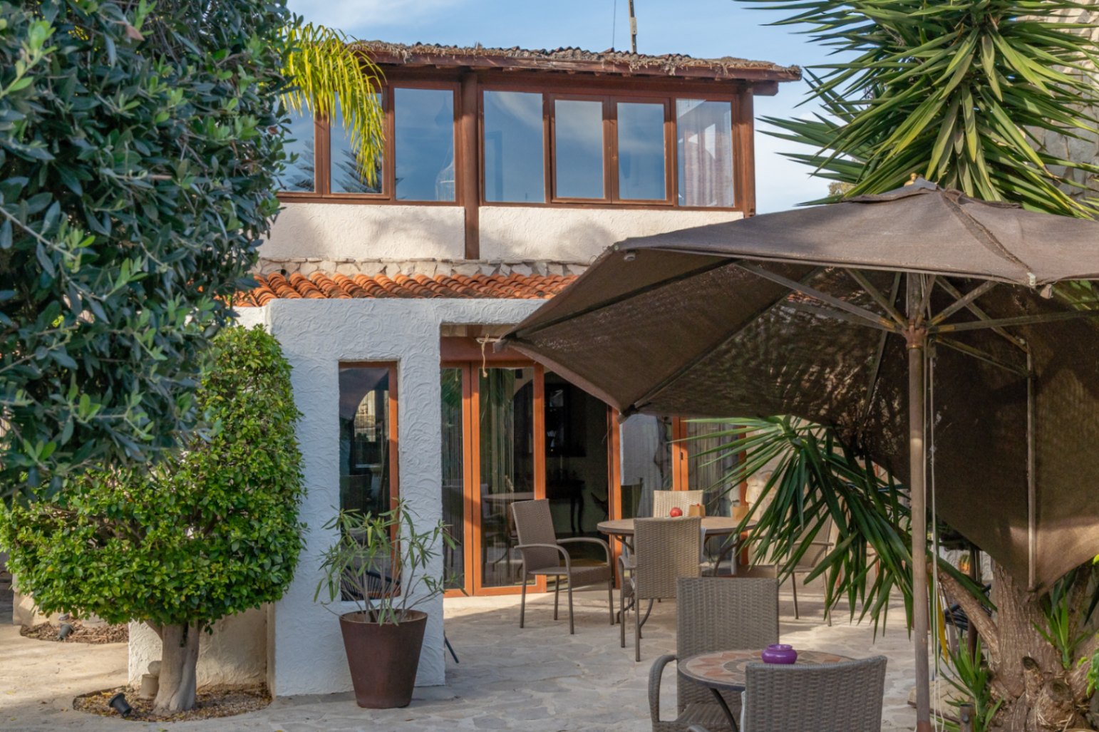Extérieur de la villa avec terrasse vitrée à l'étage, espace d'été pavé en pierre naturelle et jardin méditerranéen soigné.