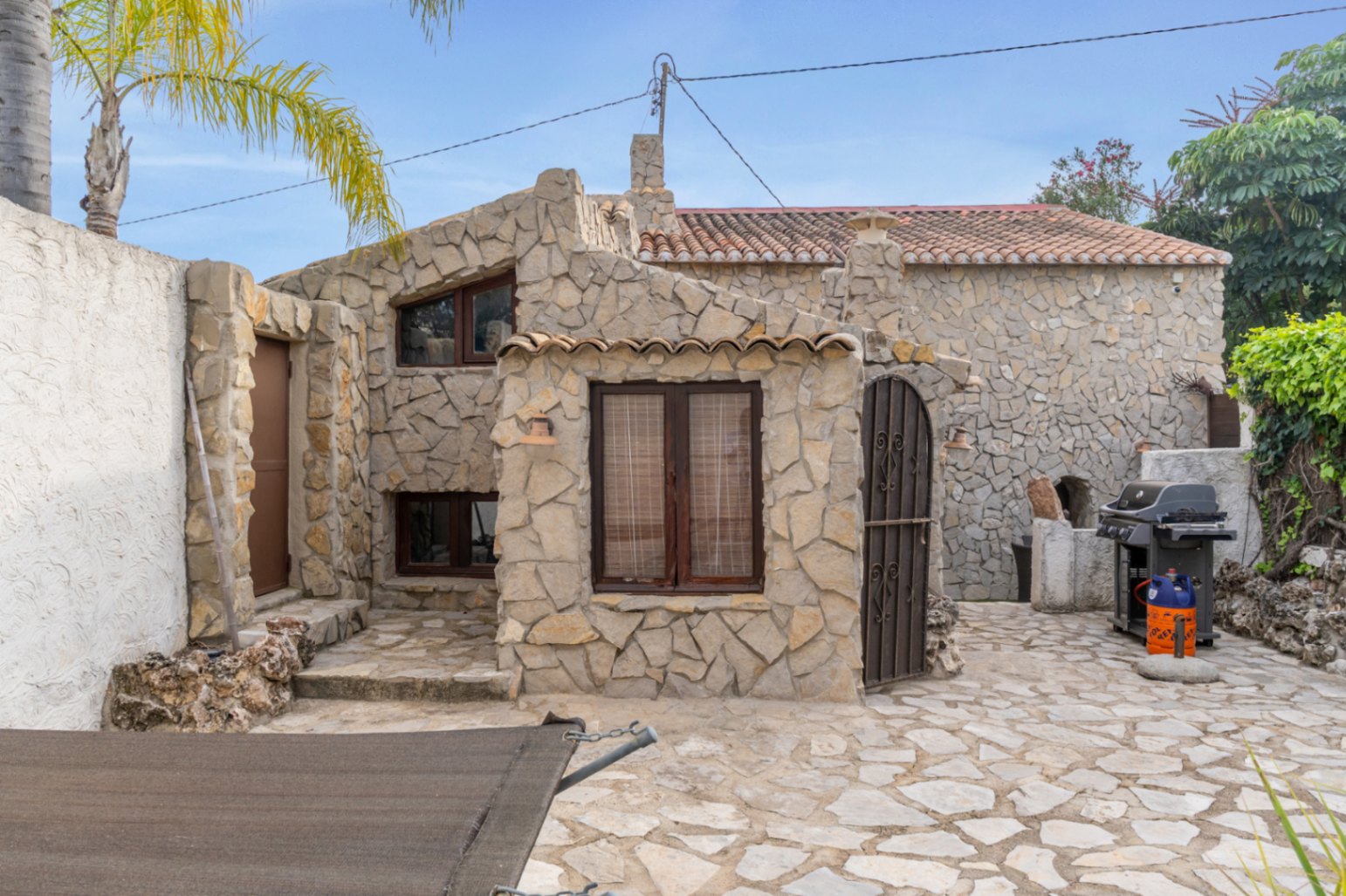 Façade en pierre naturelle avec menuiseries en bois sombre et patio pavé. L'espace extérieur comprend un barbecue et une architecture méditerranéenne rustique.