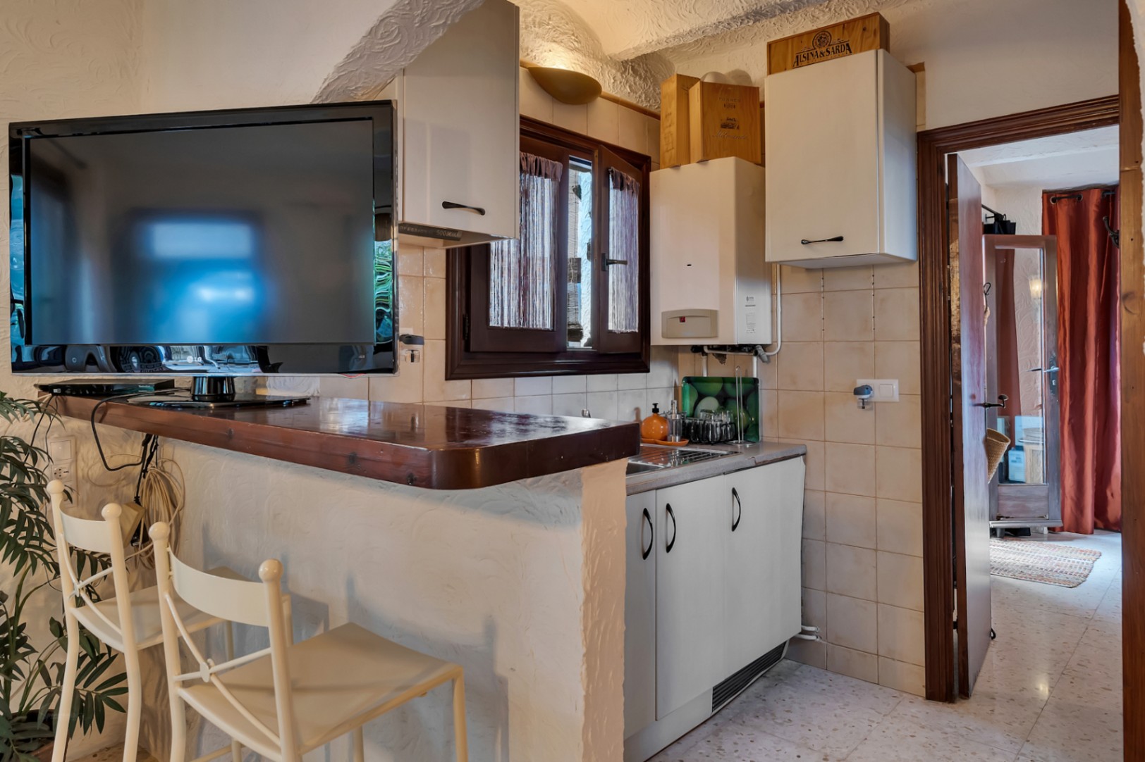 Kitchenette compacte avec bar en bois, rangements blancs et téléviseur. L'espace se distingue par ses murs en plâtre texturé et son aménagement fonctionnel.