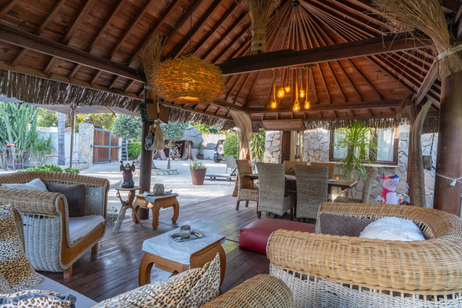 Pergola de style balinais avec charpente en bois apparente, salon de jardin en osier et espace dînatoire sur une terrasse en bois sombre.