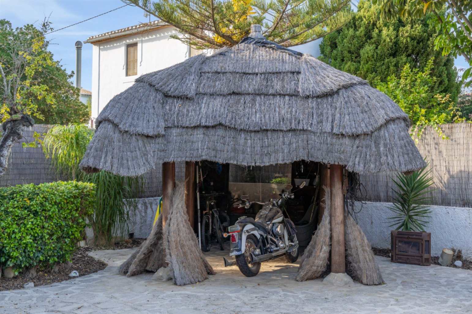 Pergola de style balinais avec toit de chaume et piliers en bois, servant d'espace de stationnement couvert pour une moto et des vélos dans le jardin méditerranéen.