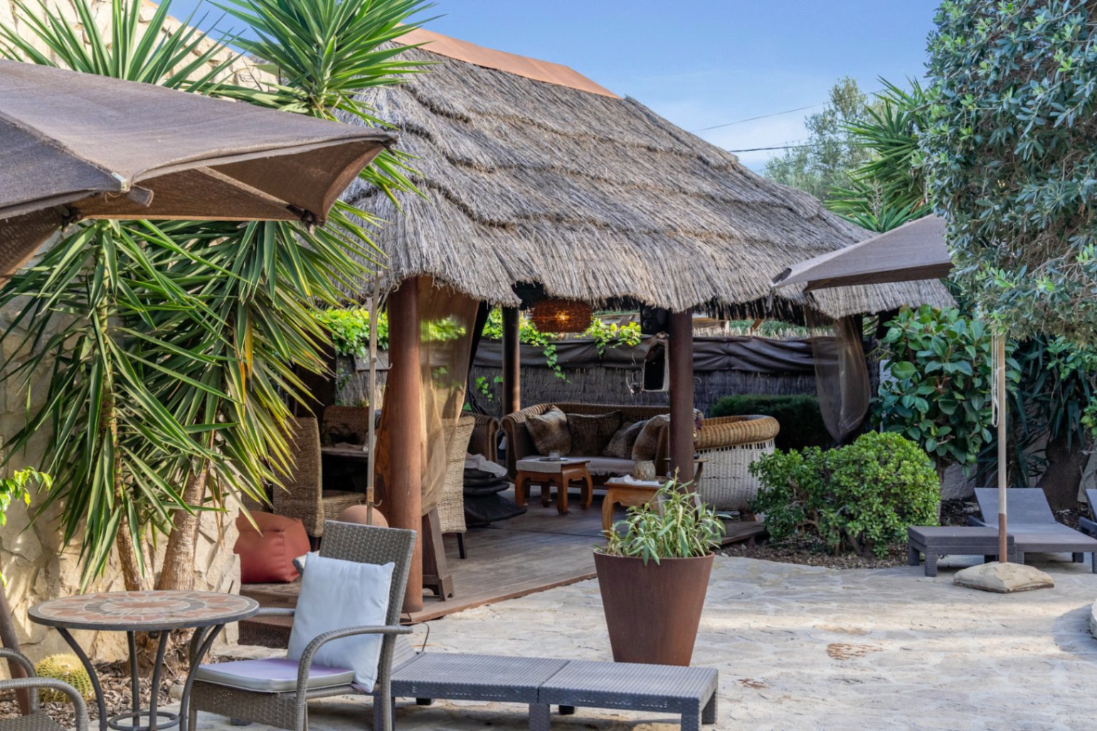 Pergola de style balinais avec toit en chaume et piliers en bois, située sur une terrasse en pierre naturelle entourée de plantes méditerranéennes et de mobilier de jardin.
