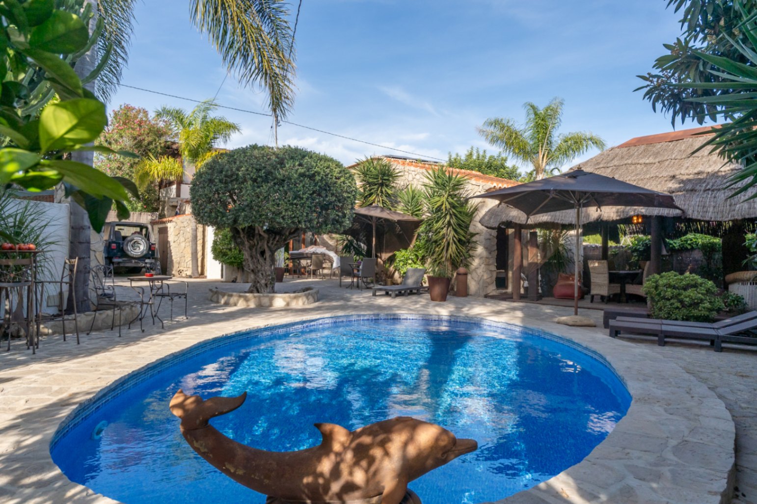 Piscine ovale entourée d'un dallage en pierre naturelle, avec une pergola de style balinais, une végétation méditerranéenne et des espaces de détente.