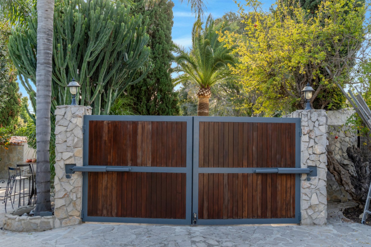 Portail d'entrée automatique avec panneaux en bois et cadre métallique, encadré par des piliers en pierre naturelle et une végétation luxuriante.