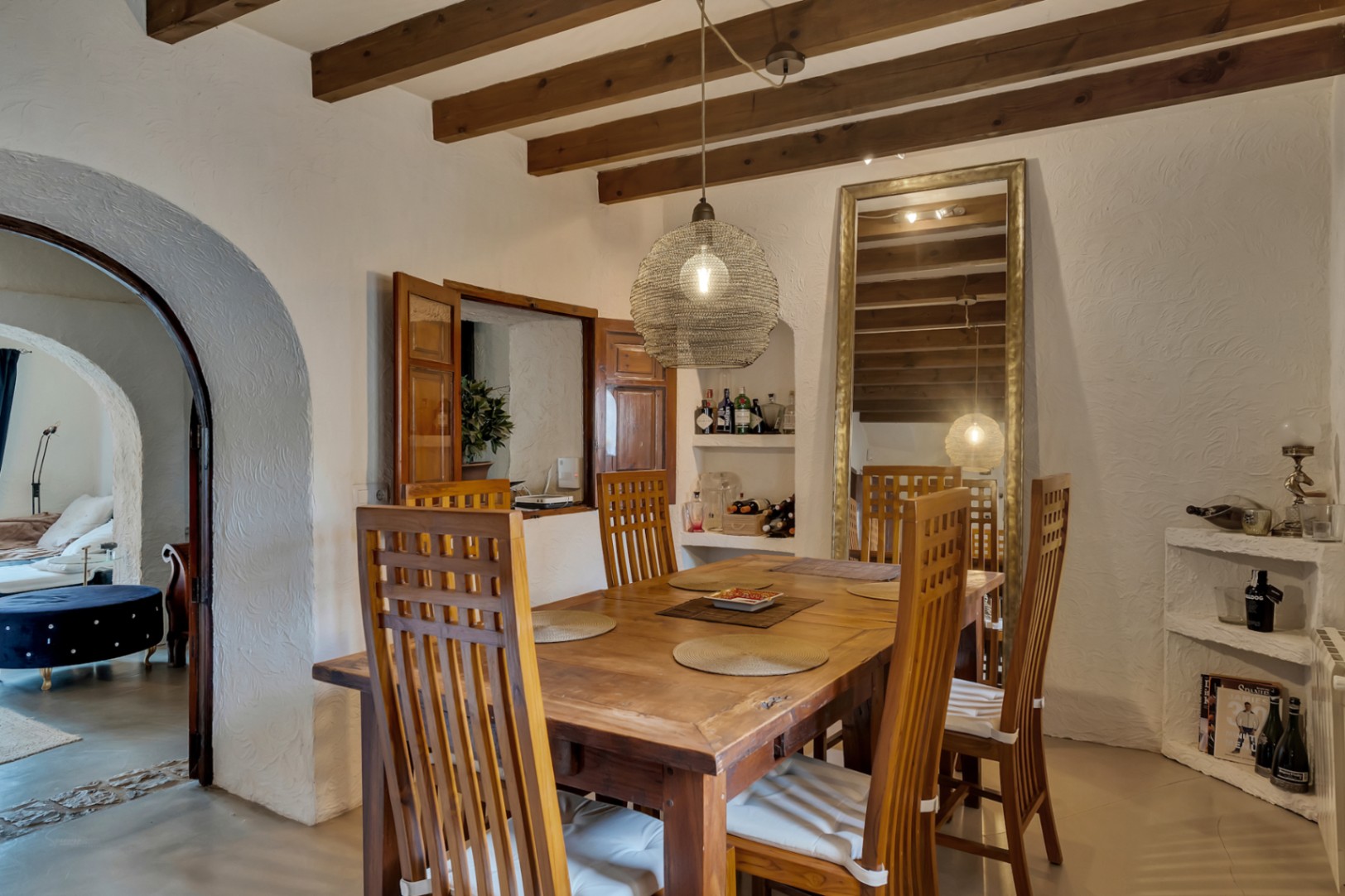 Salle à manger rustique avec poutres apparentes, table en bois massif et chaises à haut dossier. Comprend un passe-plat avec volets en bois et une arche menant aux autres pièces.