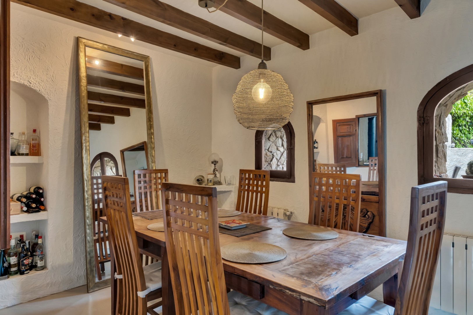 Salle à manger rustique avec poutres apparentes, table massive et chaises à haut dossier. Comprend des niches murales, de grands miroirs et une suspension en fibres naturelles.