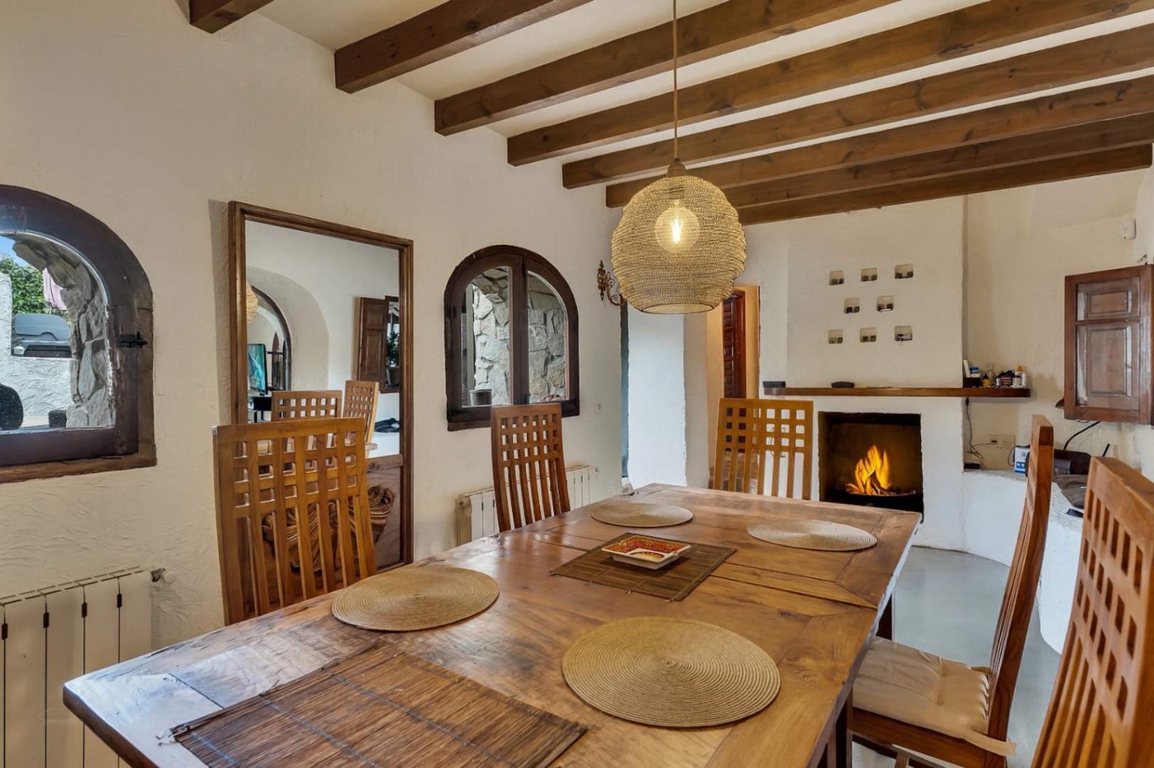 Salle à manger rustique avec poutres en bois apparentes, table massive et cheminée. Les fenêtres cintrées et la suspension en fibres naturelles créent une ambiance chaleureuse.