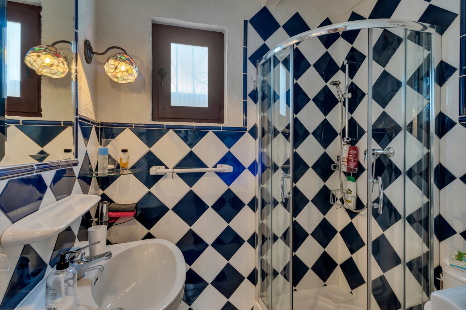 Salle de bain avec carrelage en damier bleu et blanc, cabine de douche en verre courbé et appliques murales de style Tiffany près d'une fenêtre en bois.