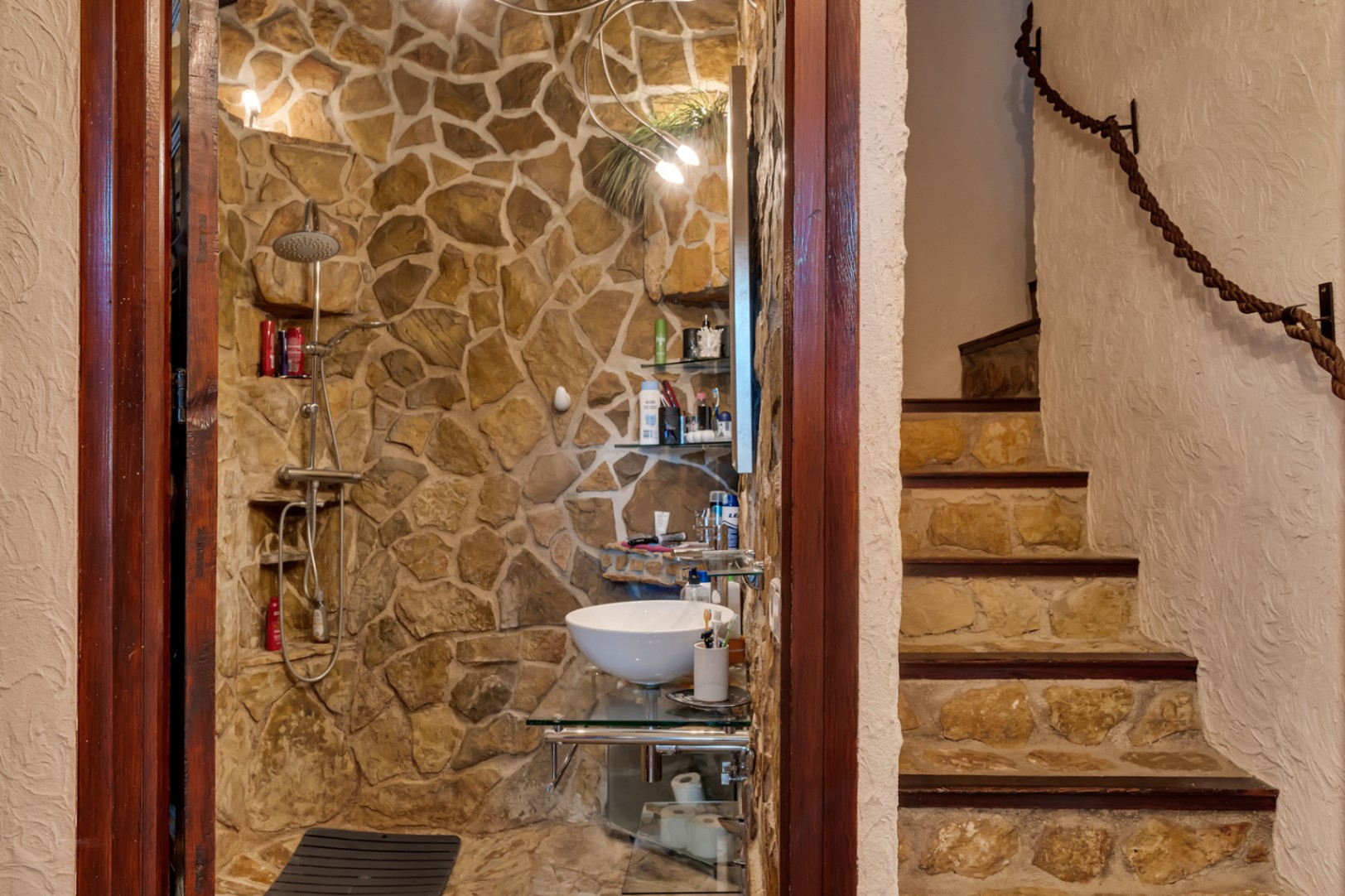 Salle de bain avec murs en pierre naturelle, vasque sur plan en verre et douche intégrée. Escalier attenant avec contremarches en pierre et rampe en corde.
