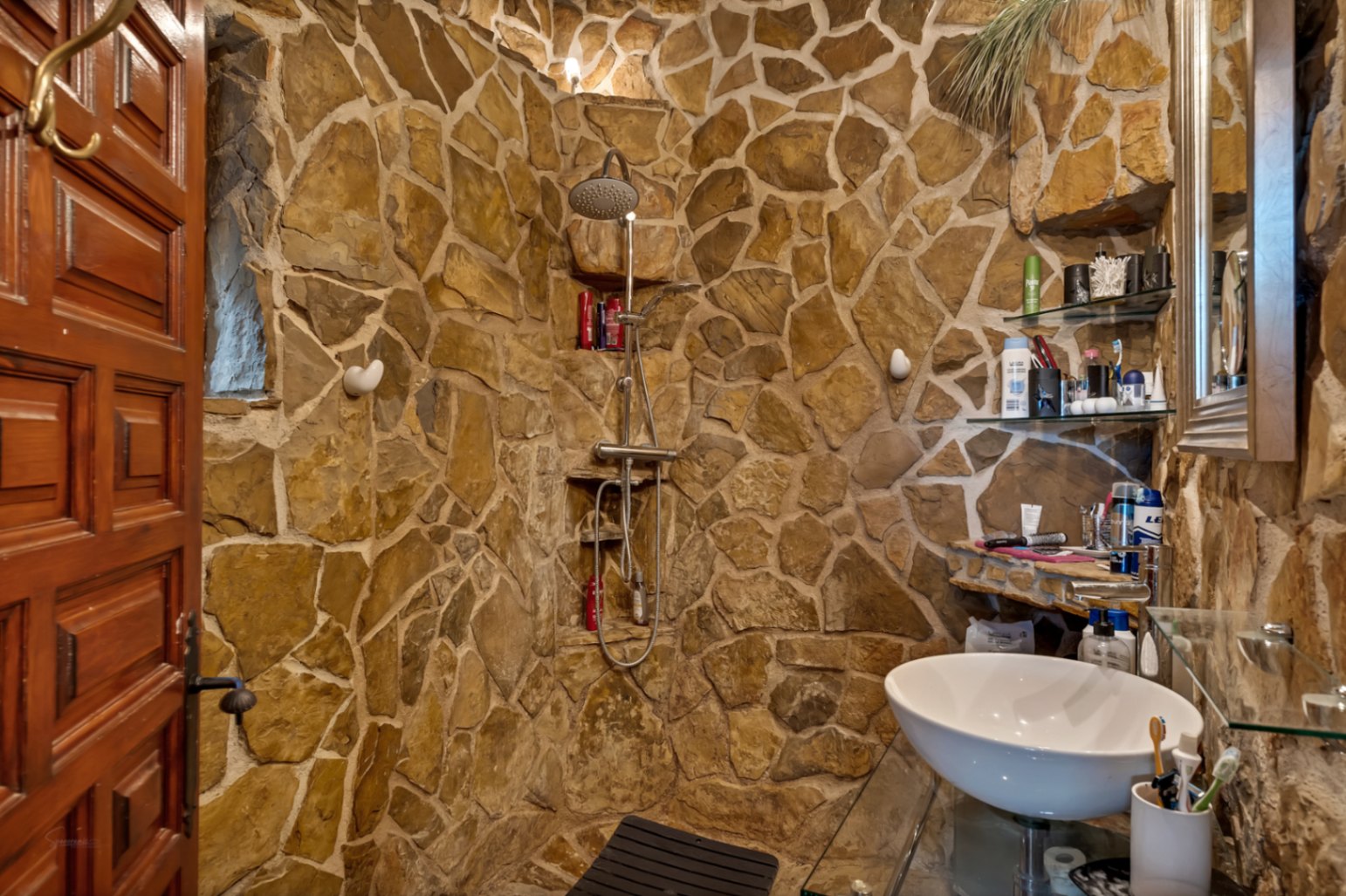 Salle de bain rustique avec murs en pierre naturelle, douche intégrée avec robinetterie chromée et vasque blanche sur un plan de travail en verre.