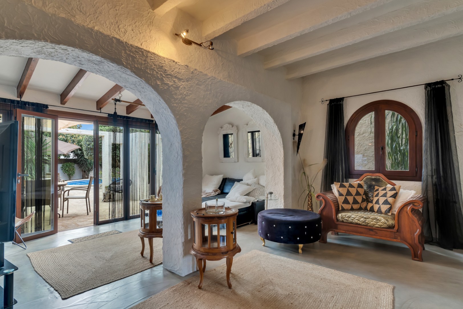 Salon aux arches traditionnelles et plafonds à poutres blanches. L'espace s'ouvre sur une naya lumineuse avec accès direct à la terrasse et à la piscine extérieure.