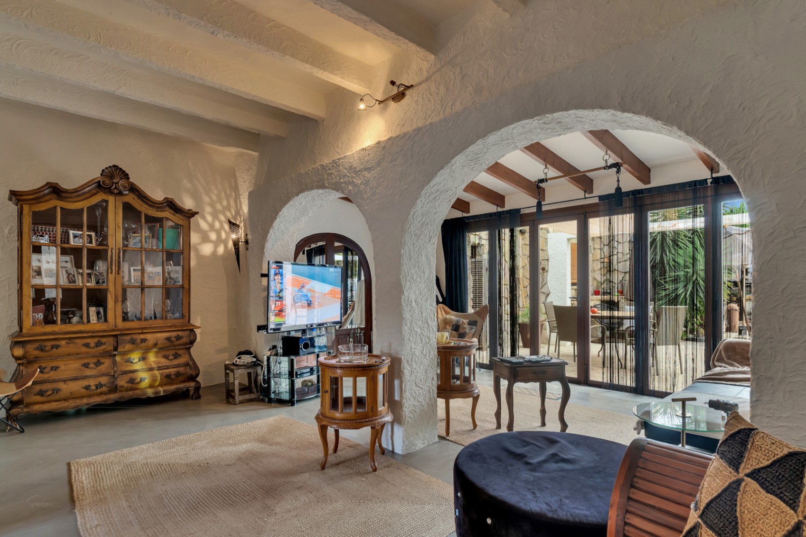 Salon intérieur avec plafonds à poutres apparentes et arches blanches texturées ouvrant sur une naya vitrée. Un buffet classique en bois sculpté complète l'espace.