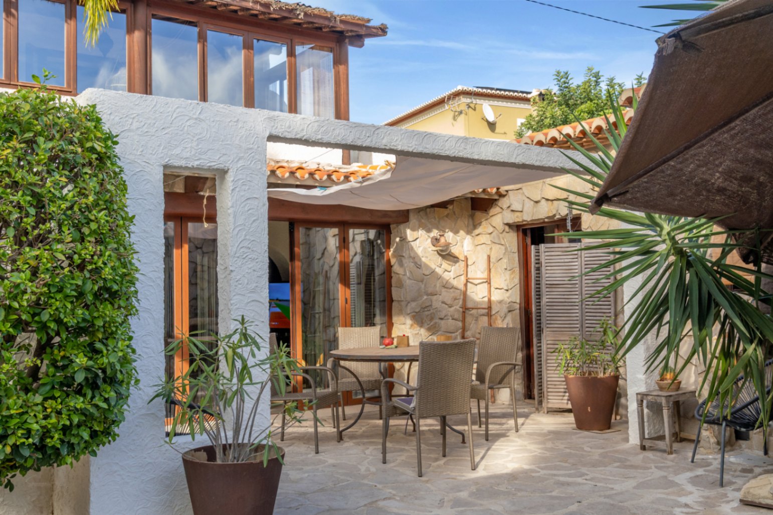 Terrasse extérieure avec sol en pierre, espace dînatoire sous une voile d'ombrage et façade alliant maçonnerie de pierre et menuiseries en bois.