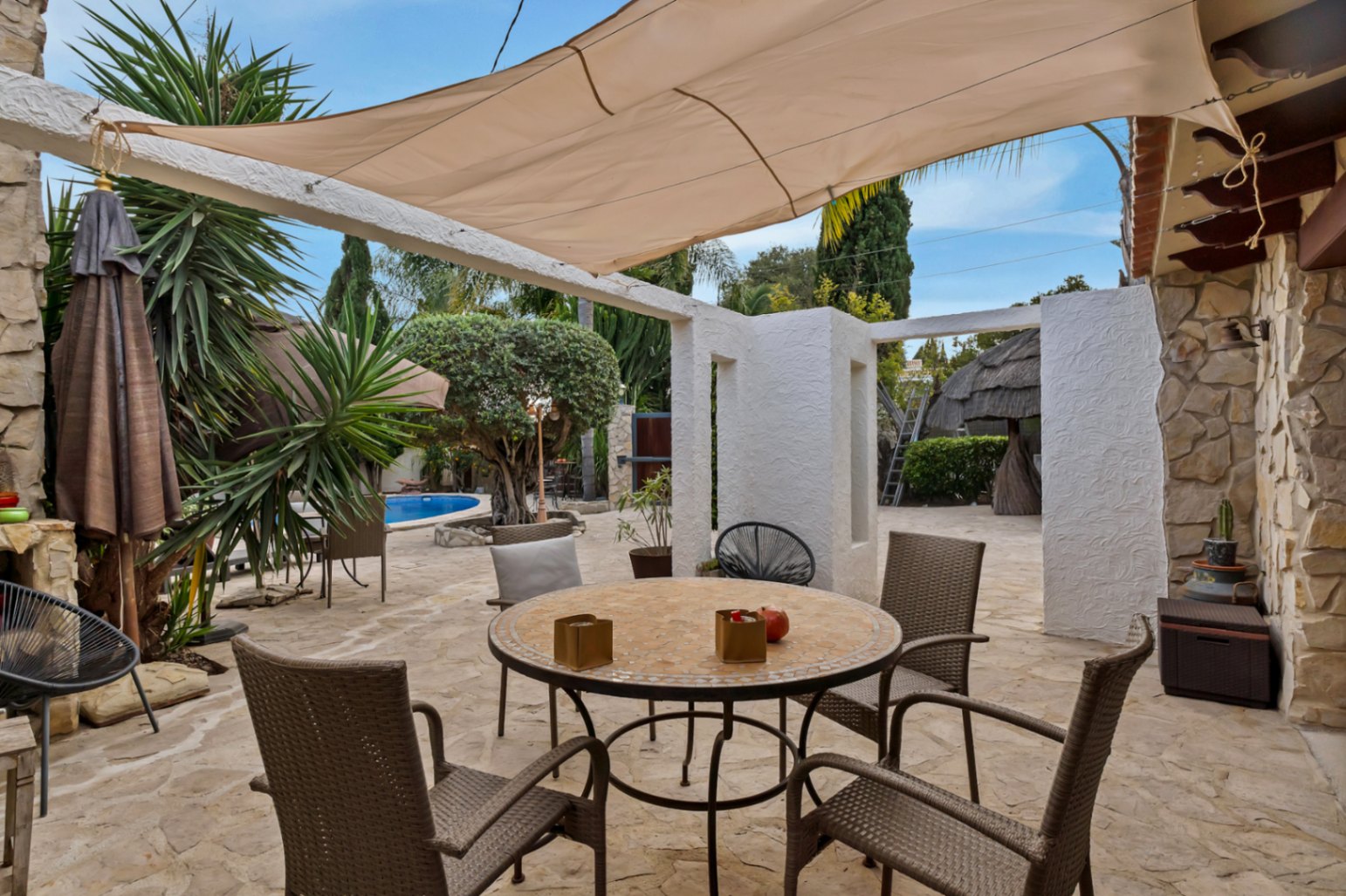 Terrasse extérieure avec sol en pierre naturelle, coin repas abrité par une voile d'ombrage et vue sur le jardin méditerranéen et la piscine.