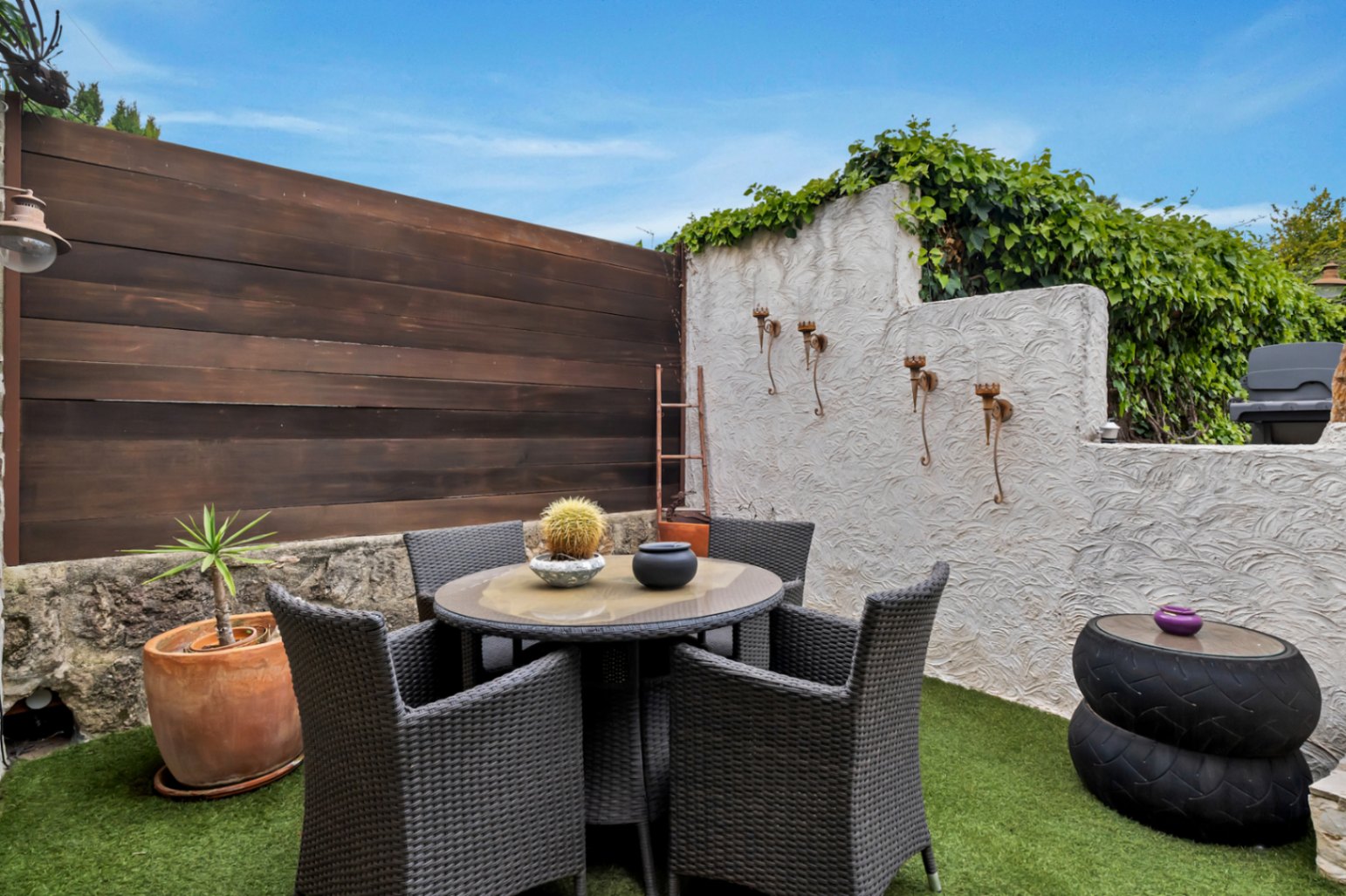 Terrasse privée avec gazon synthétique, salle à manger d'extérieur en résine tressée et murs mêlant bois, pierre et crépi blanc texturé.