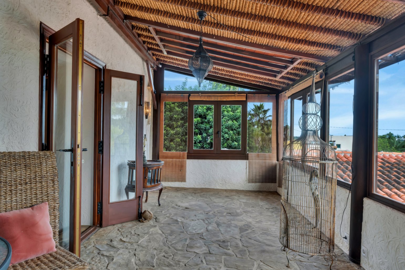 Terrasse vitrée avec sol en pierre naturelle, plafond en roseau avec poutres en bois et lanterne marocaine, offrant une vue dégagée sur la végétation.