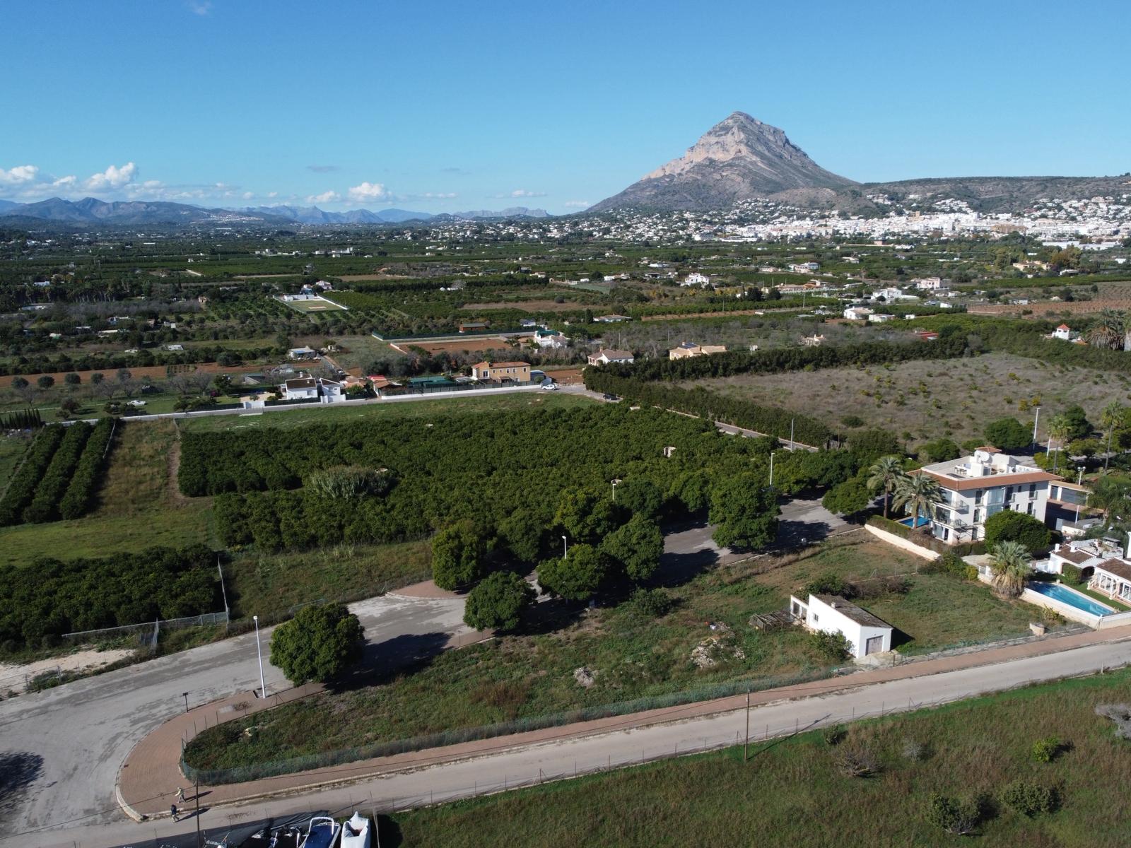 Vue aérienne du terrain à bâtir avec accès goudronné, entouré de zones agricoles verdoyantes et offrant une vue dégagée sur le mont Montgó.