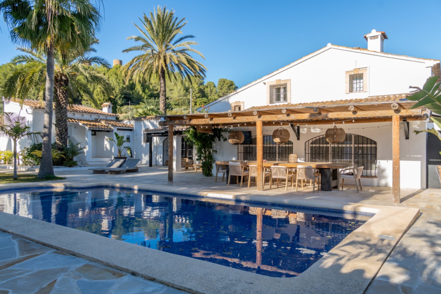 Espace extérieur avec piscine rectangulaire et pergola en bois. La salle à manger d'été dispose d'une grande table, de suspensions en osier et d'un cadre méditerranéen authentique.