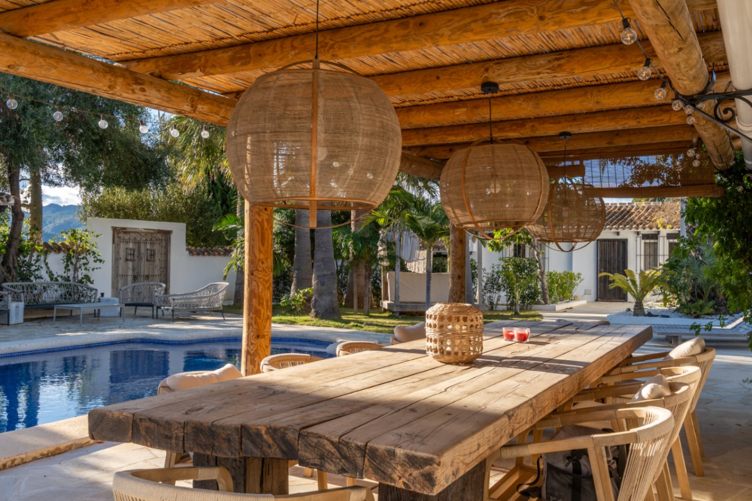 Salle à manger extérieure sous une pergola en bois et canisse, avec table rustique, suspensions en fibres naturelles et vue sur la piscine et le jardin méditerranéen.