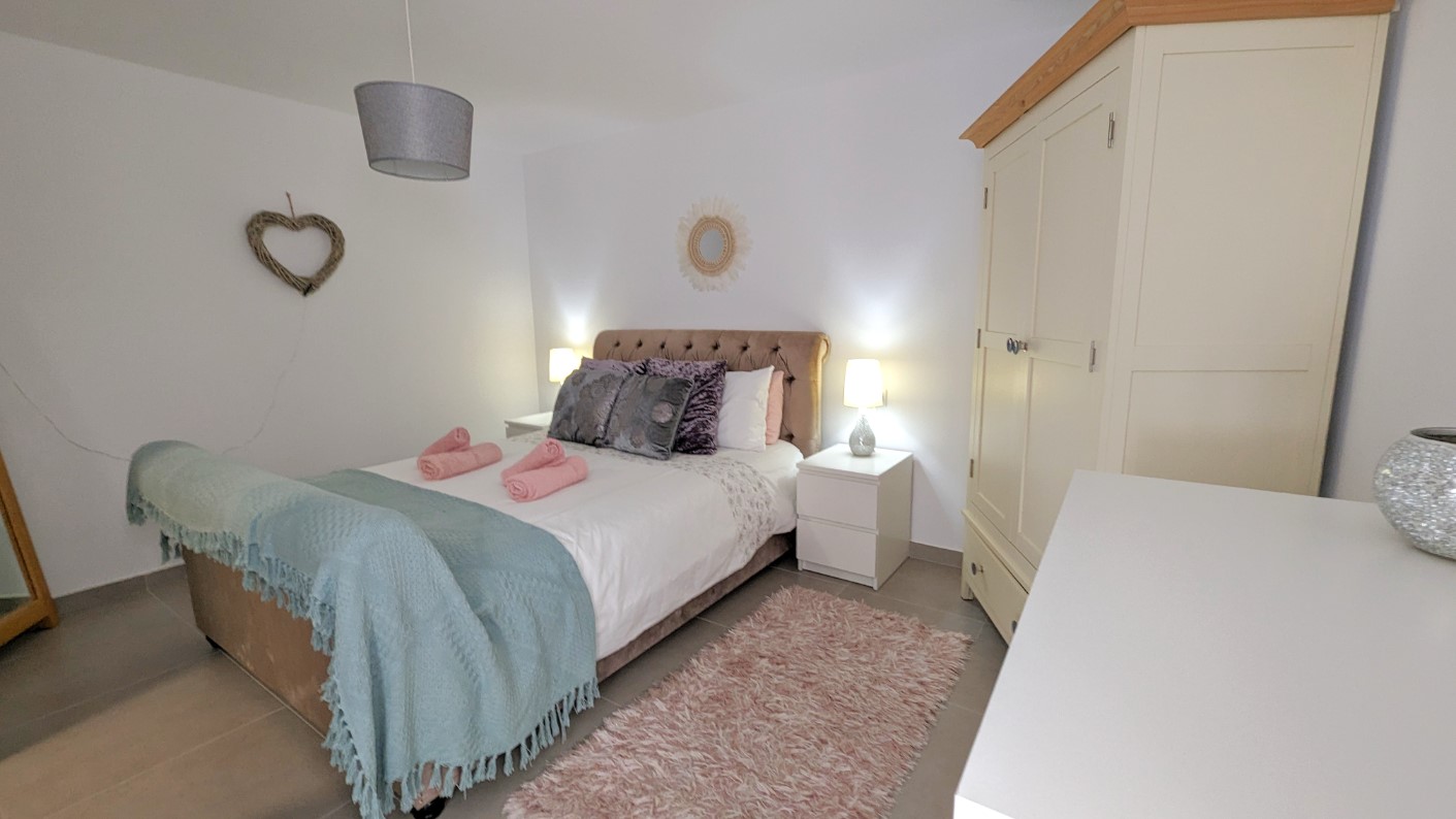 Chambre à coucher avec un lit double à tête de lit capitonnée, des tables de chevet blanches et une armoire crème. Le sol est en carrelage gris clair avec des murs blancs.