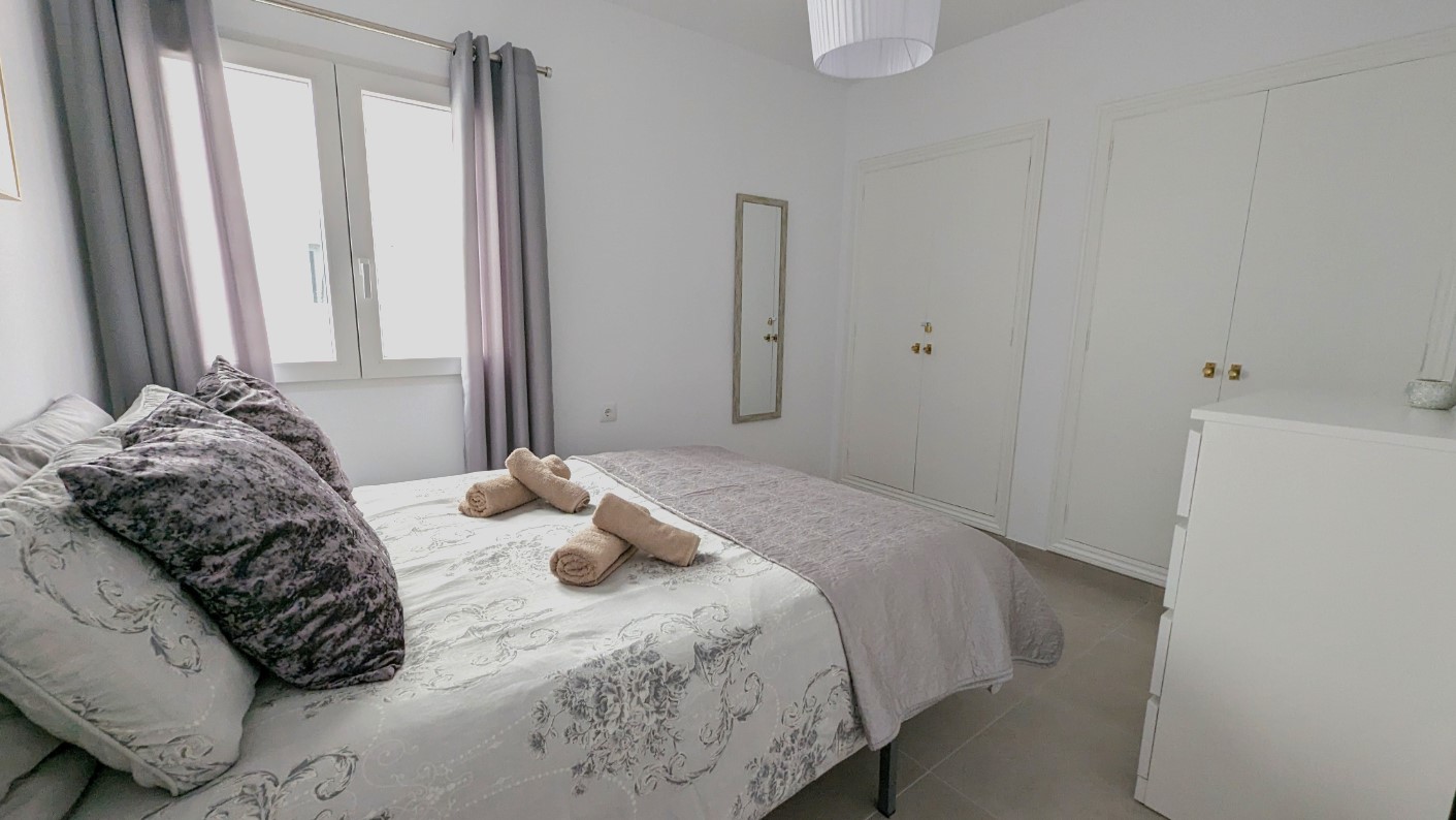 Chambre double avec placards intégrés blancs, miroir mural et sol en carrelage. La pièce bénéficie d'une lumière naturelle constante et d'une décoration aux tons neutres.