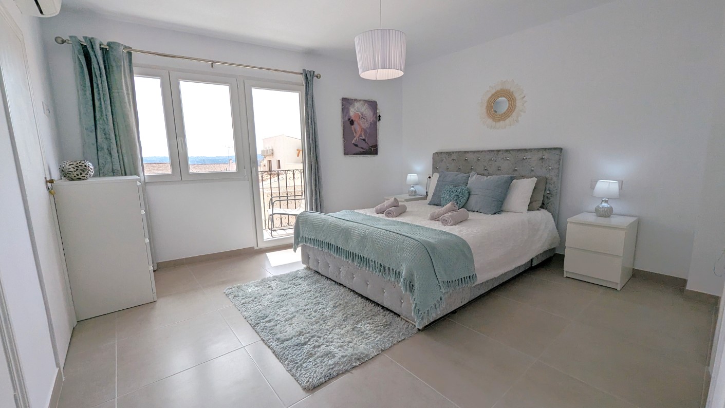 Chambre principale avec tête de lit capitonnée, accès à une terrasse privée par des portes vitrées et mobilier blanc aux lignes épurées.