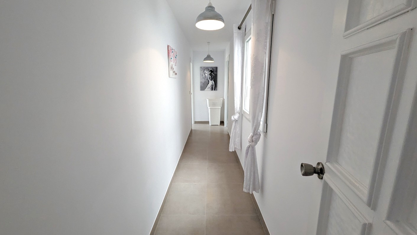 Couloir lumineux avec murs blancs, carrelage au sol, suspensions de style industriel et fenêtres avec rideaux blancs noués.