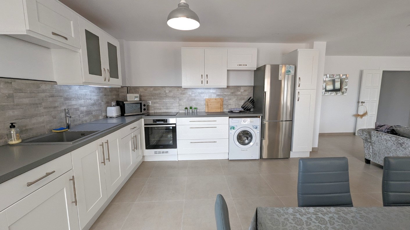 Cuisine moderne ouverte avec placards blancs de style shaker, crédence en carreaux gris effet pierre et appareils électroménagers intégrés en acier inoxydable.