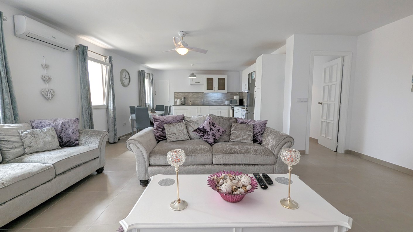 Espace de vie ouvert comprenant des canapés en velours gris, une table basse blanche et un ventilateur de plafond. La pièce intègre un coin repas et une cuisine blanche moderne.