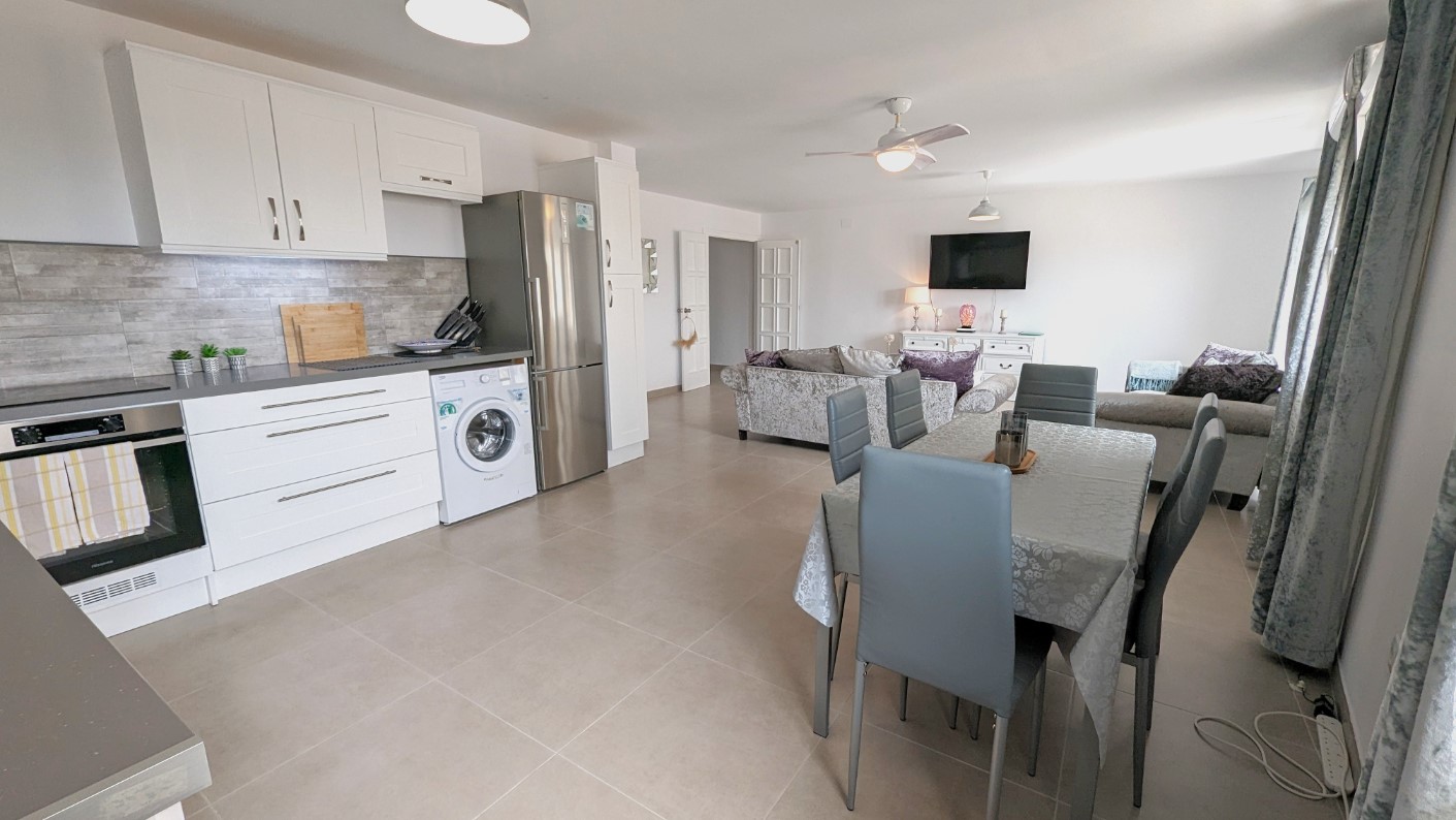 Espace de vie ouvert comprenant une cuisine blanche moderne équipée, un coin repas avec chaises grises et un salon avec ventilateur de plafond et carrelage clair.