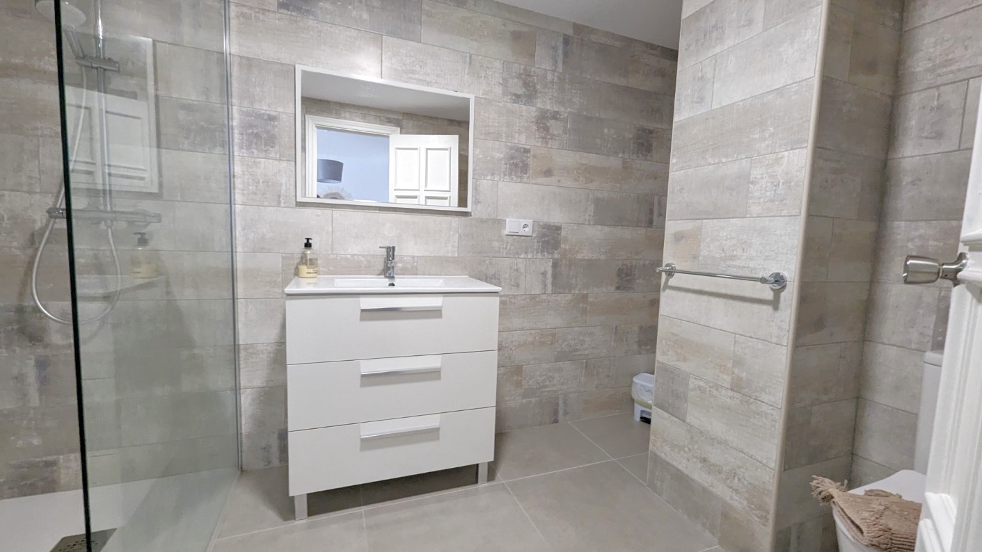 Salle de bain moderne avec meuble vasque blanc à trois tiroirs, douche à l'italienne avec paroi vitrée et carrelage mural texturé aux tons naturels.