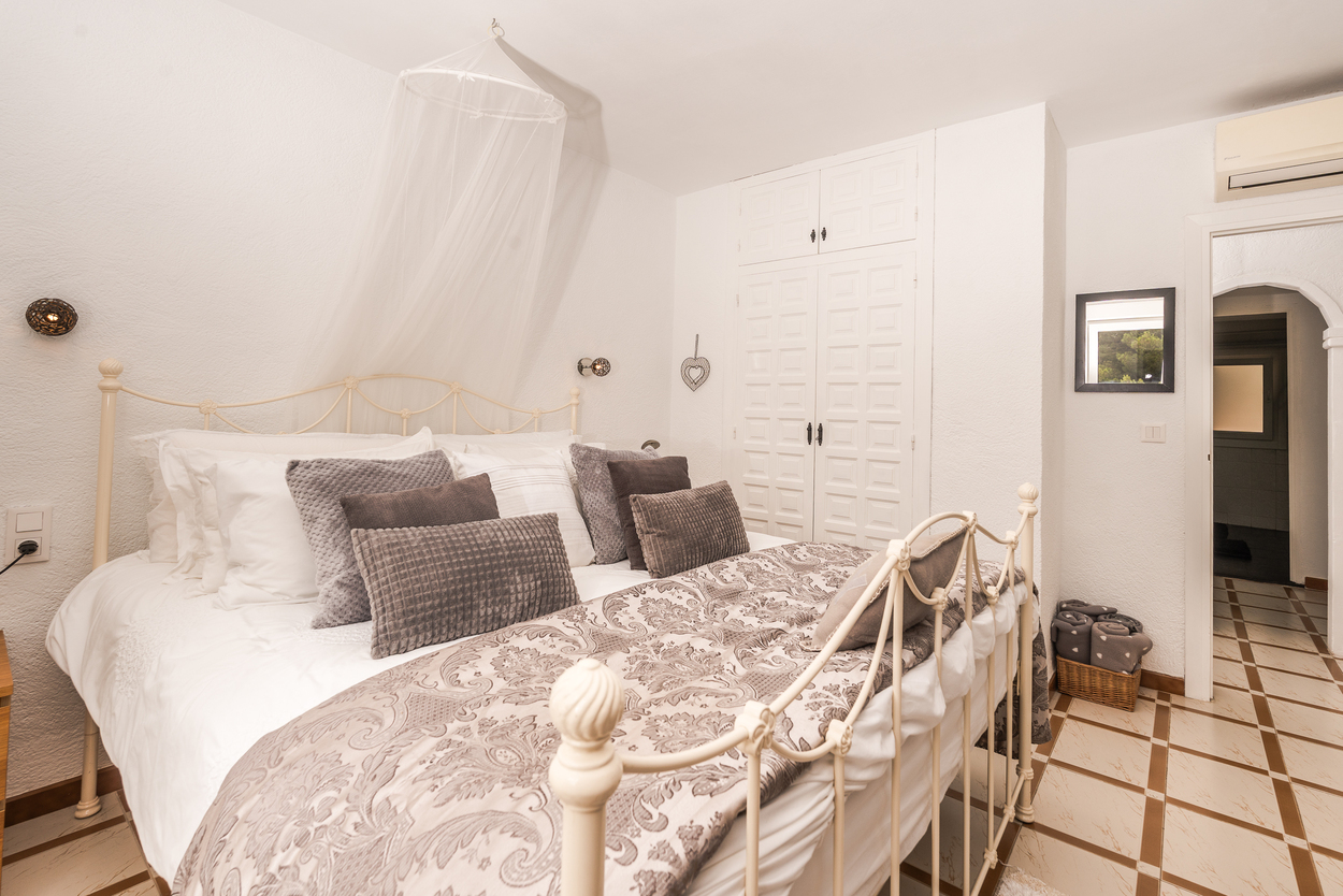 Chambre avec lit en fer forgé blanc, moustiquaire en baldaquin et placards intégrés. Sol en carrelage et climatisation installée au-dessus de la porte voûtée.